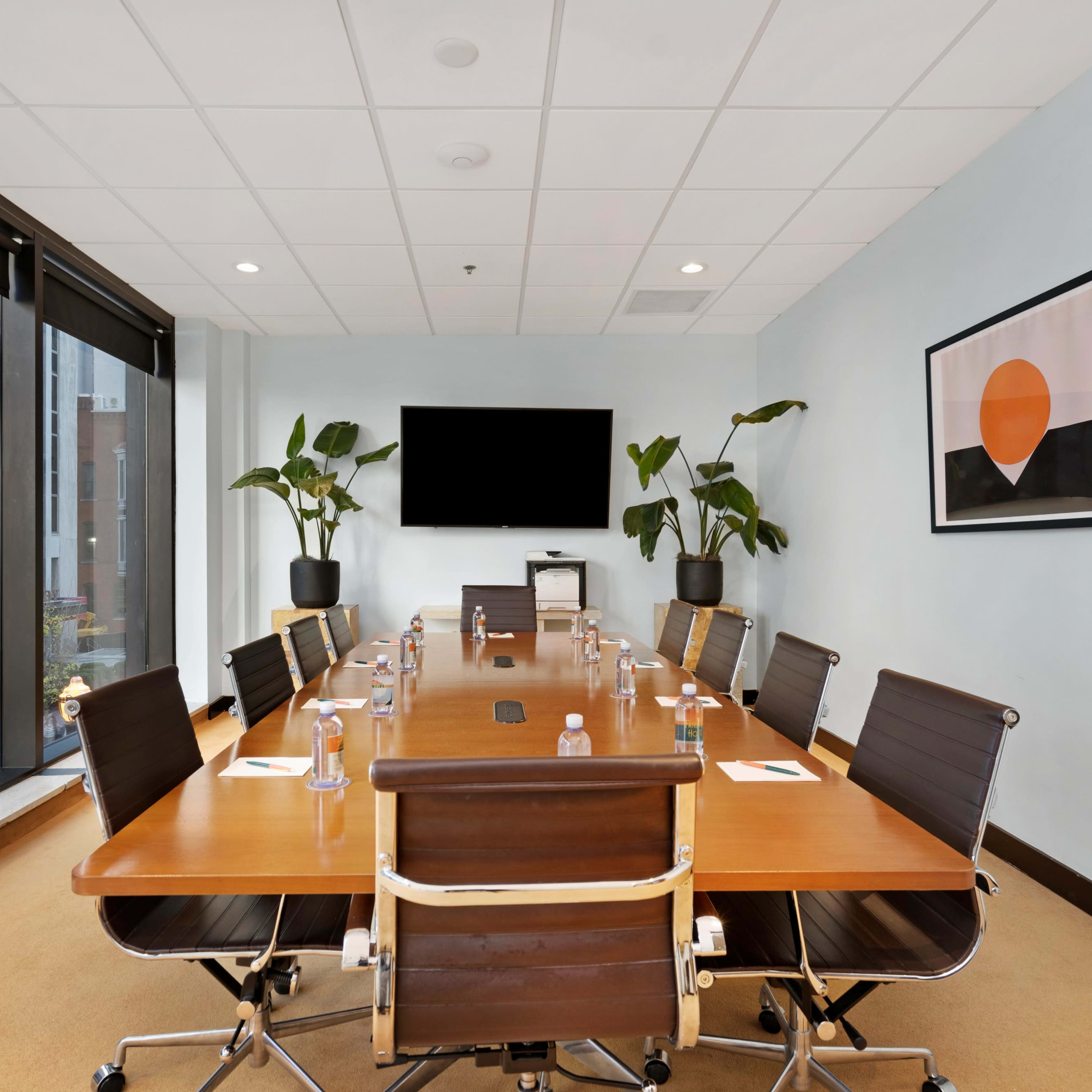 A modern conference room with a long wooden table, black leather chairs, a wall-mounted TV, and large windows overlooking a city street.