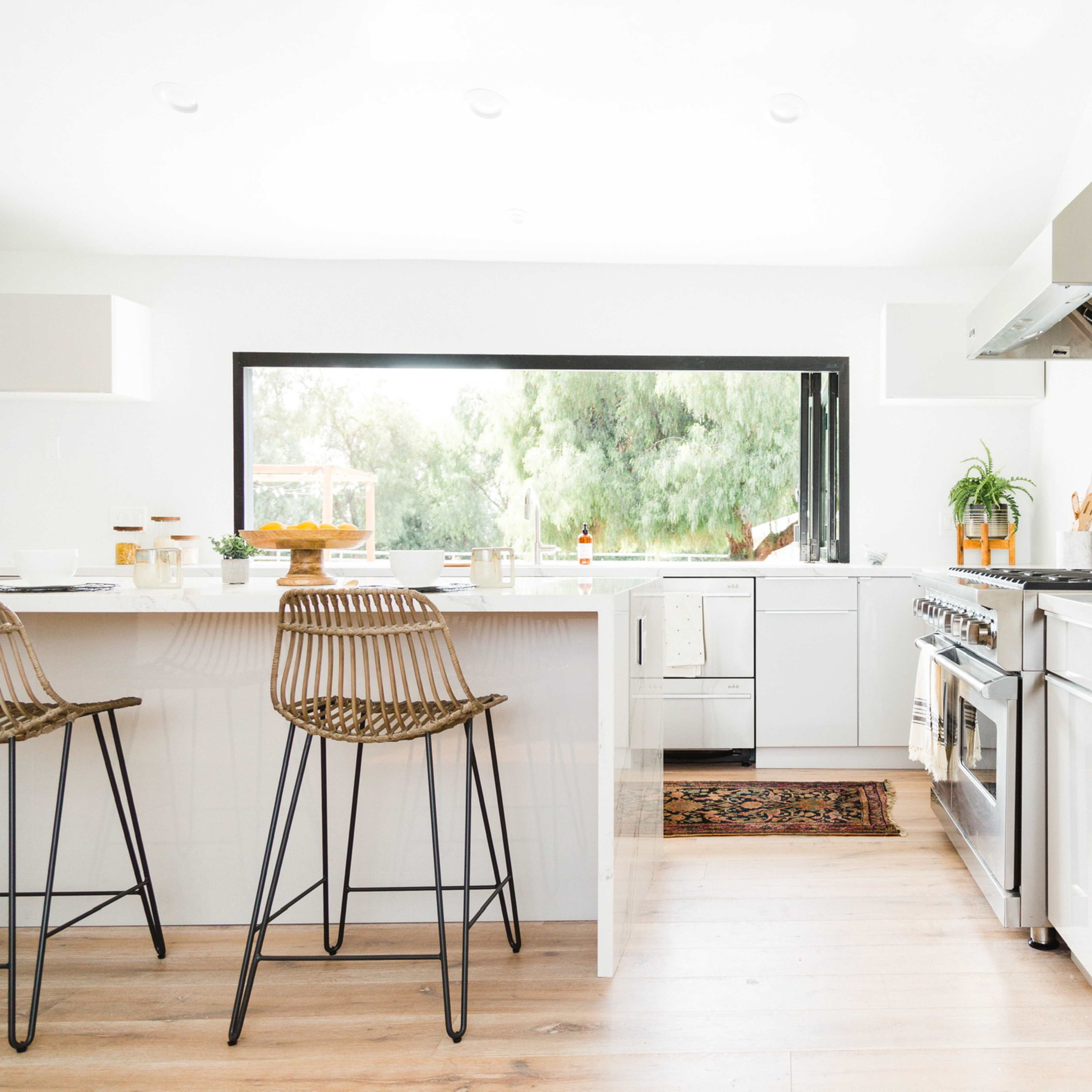 A modern kitchen features a large island with two bar stools, stainless steel appliances, and a window overlooking greenery.