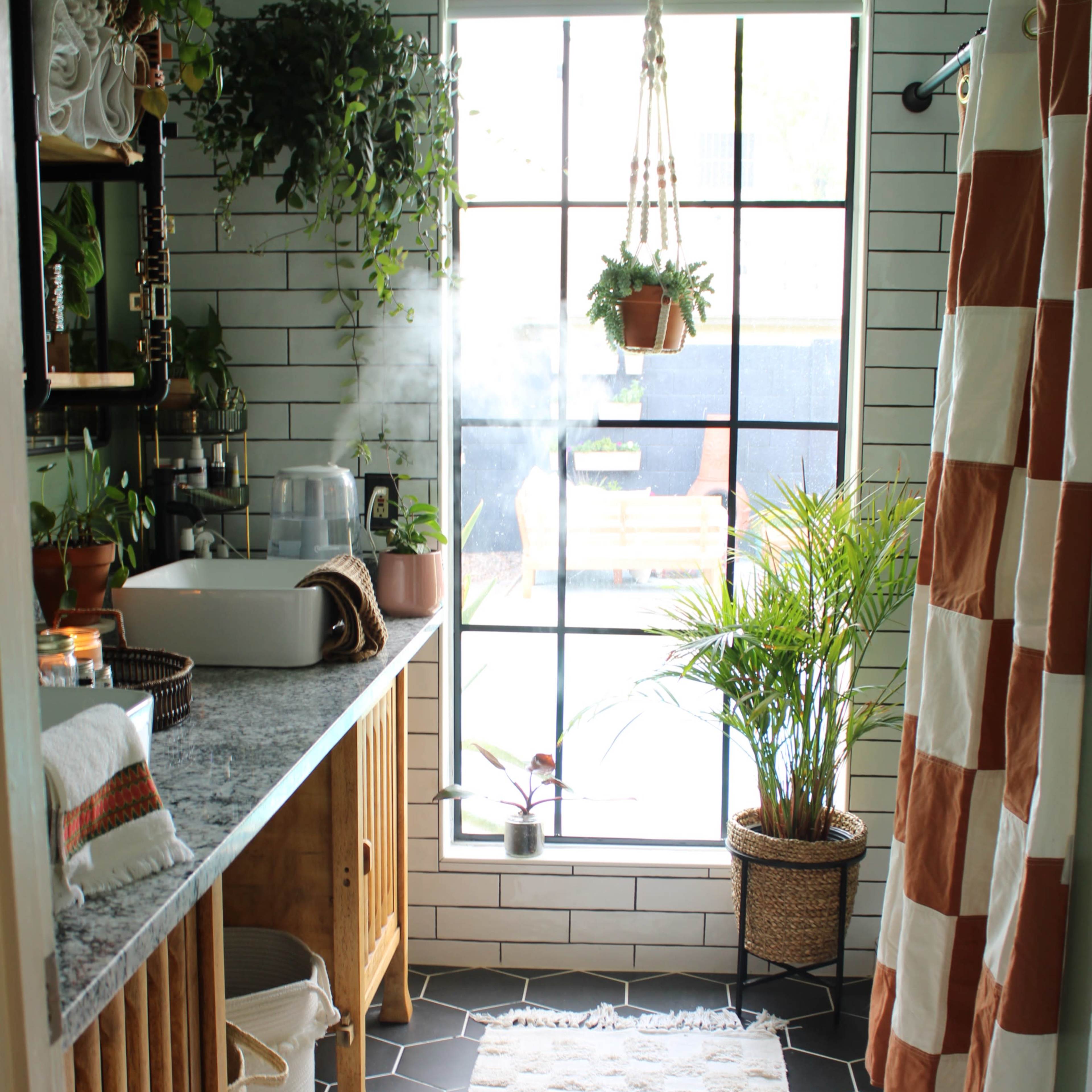 A bathroom with a large window, a marble countertop, wooden cabinetry, plants, and a patterned shower curtain.