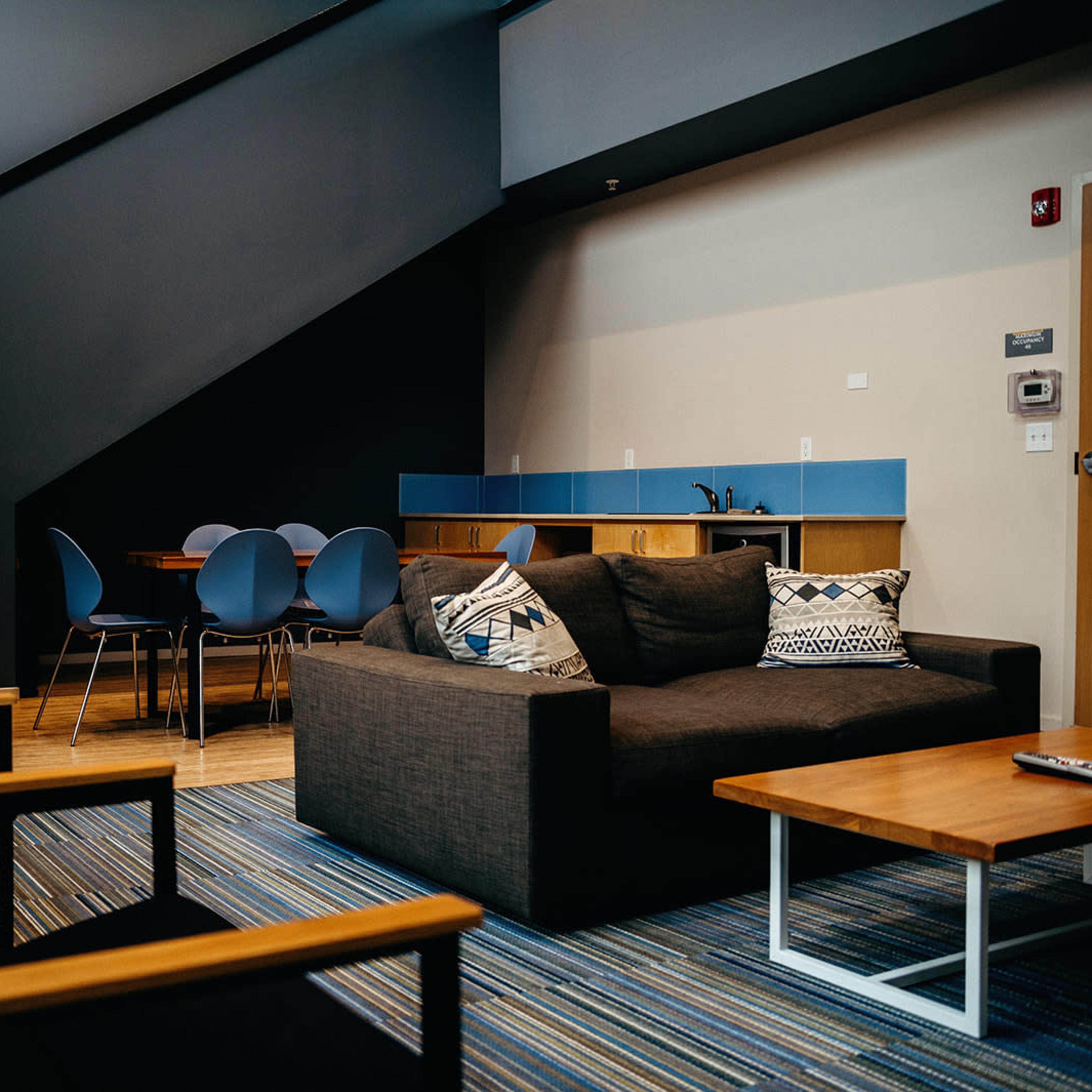 A modern lounge area features a brown sofa, wooden coffee tables, and a dining space with blue chairs adjacent to a staircase.