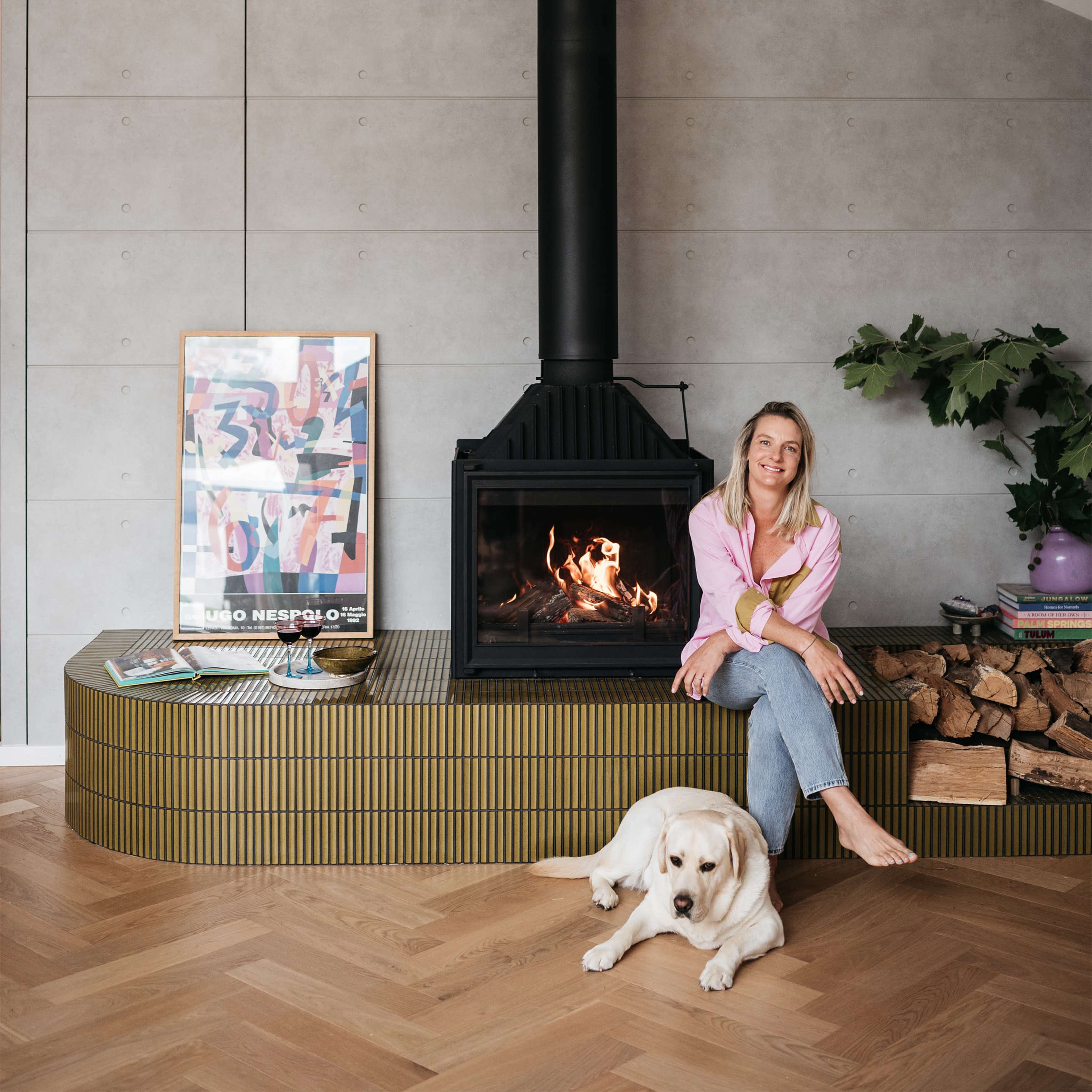 A woman sits on a curved, yellow-striped bench beside a black wood-burning stove, with a golden retriever lying on the floor in front of her.