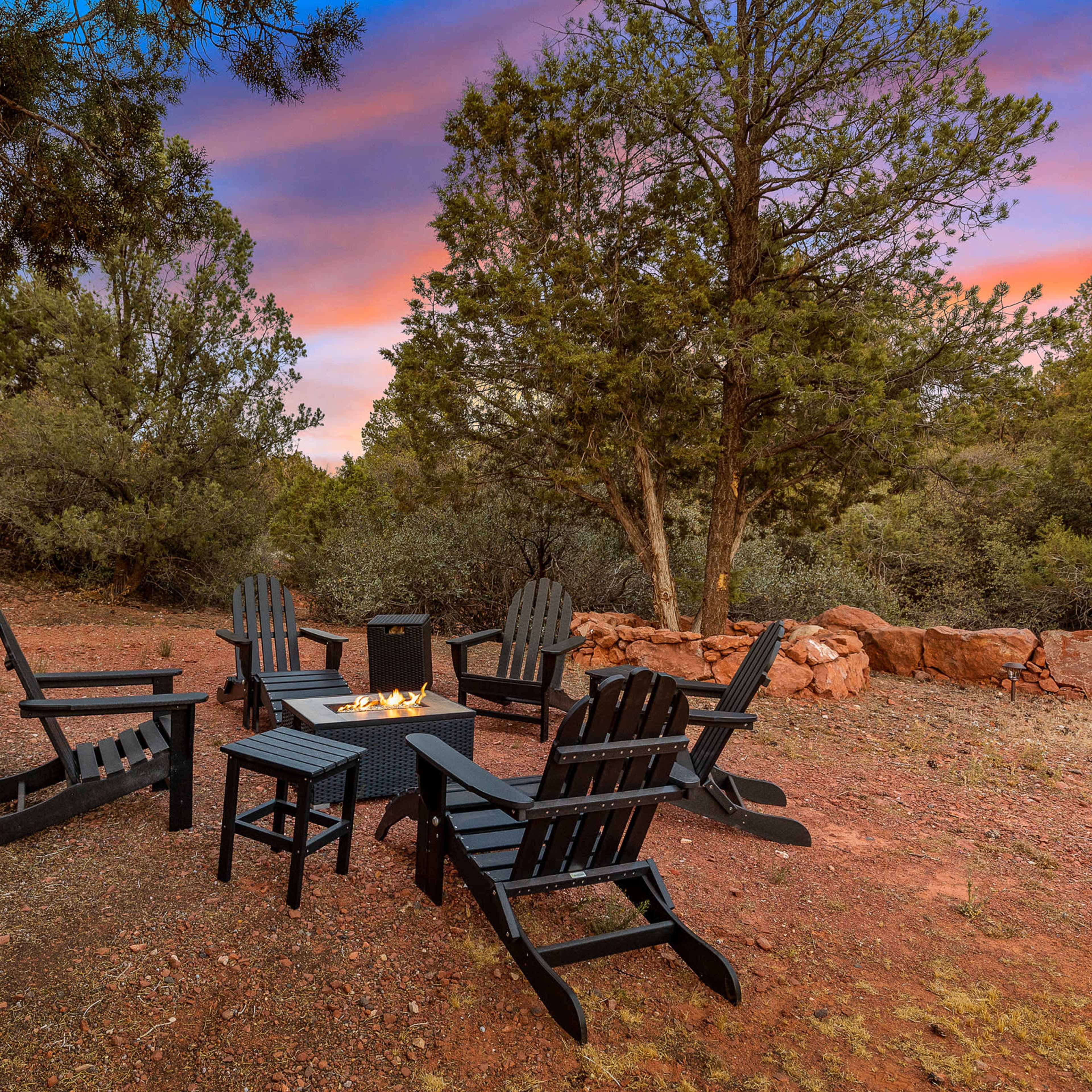 A circle of black Adirondack chairs surrounds a central fire pit in a wooded outdoor setting at dusk.