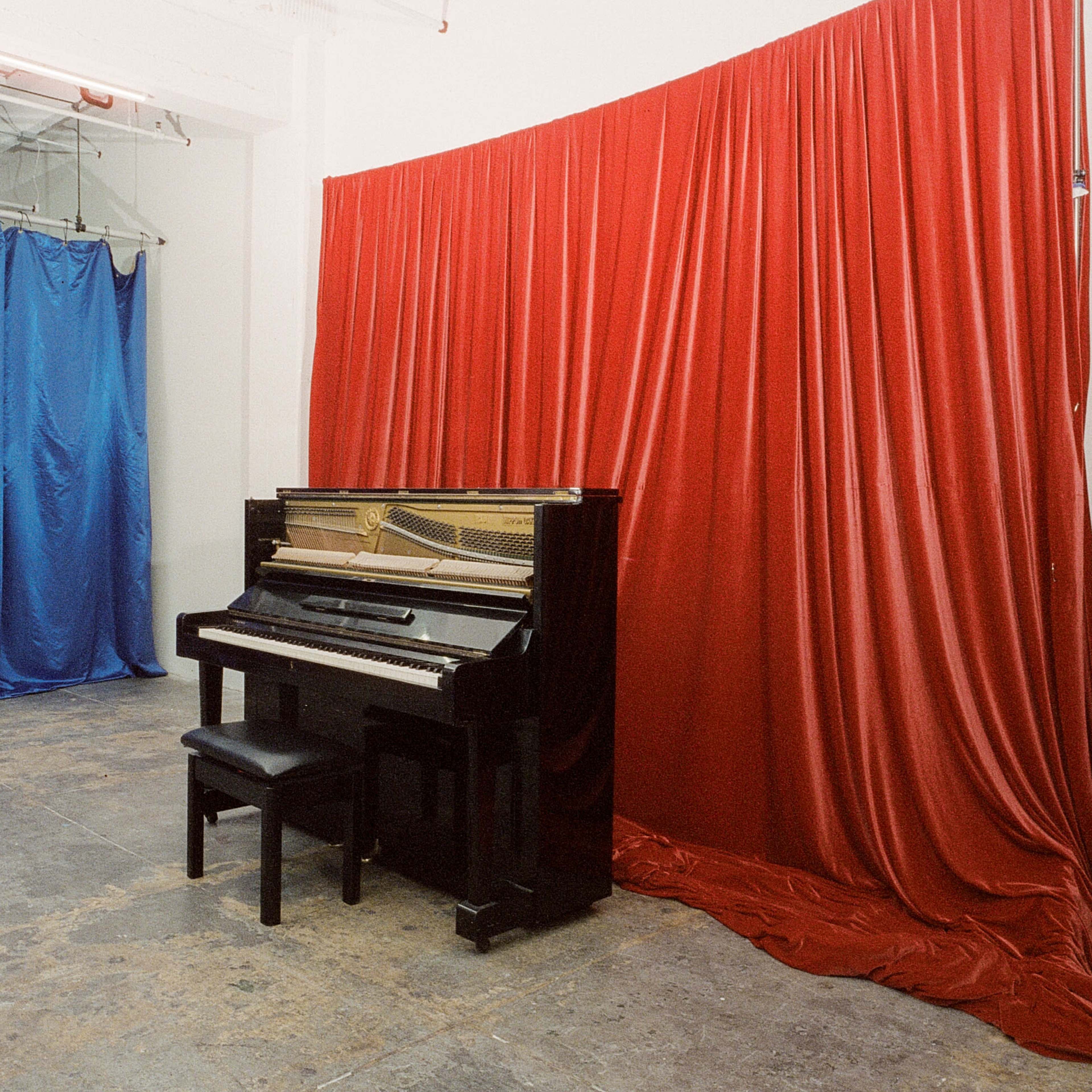 A black piano with a matching stool positioned against a red curtain, with blue drapes in the background.