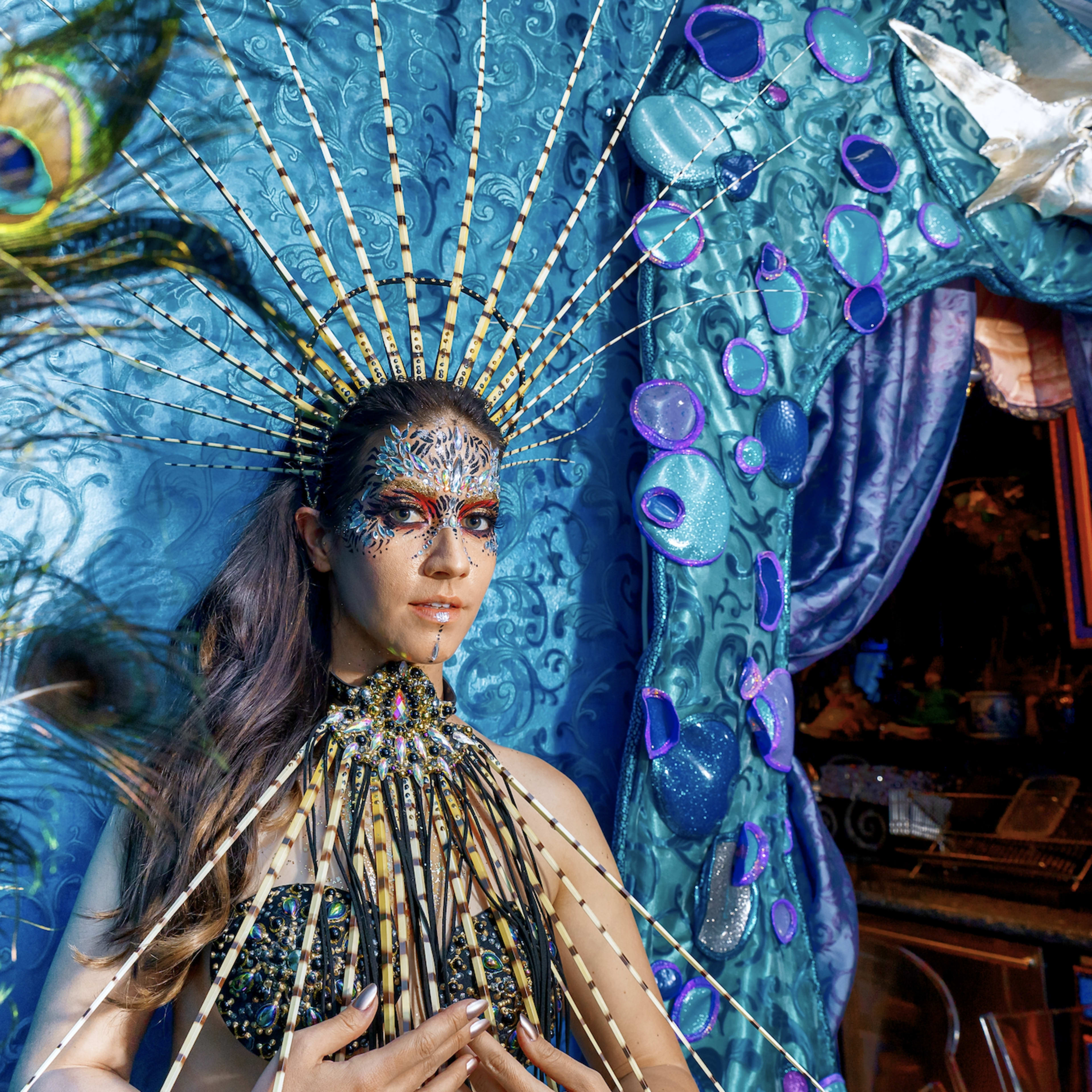 A woman with ornate body paint and elaborate accessories poses against a vibrant, decorated backdrop.