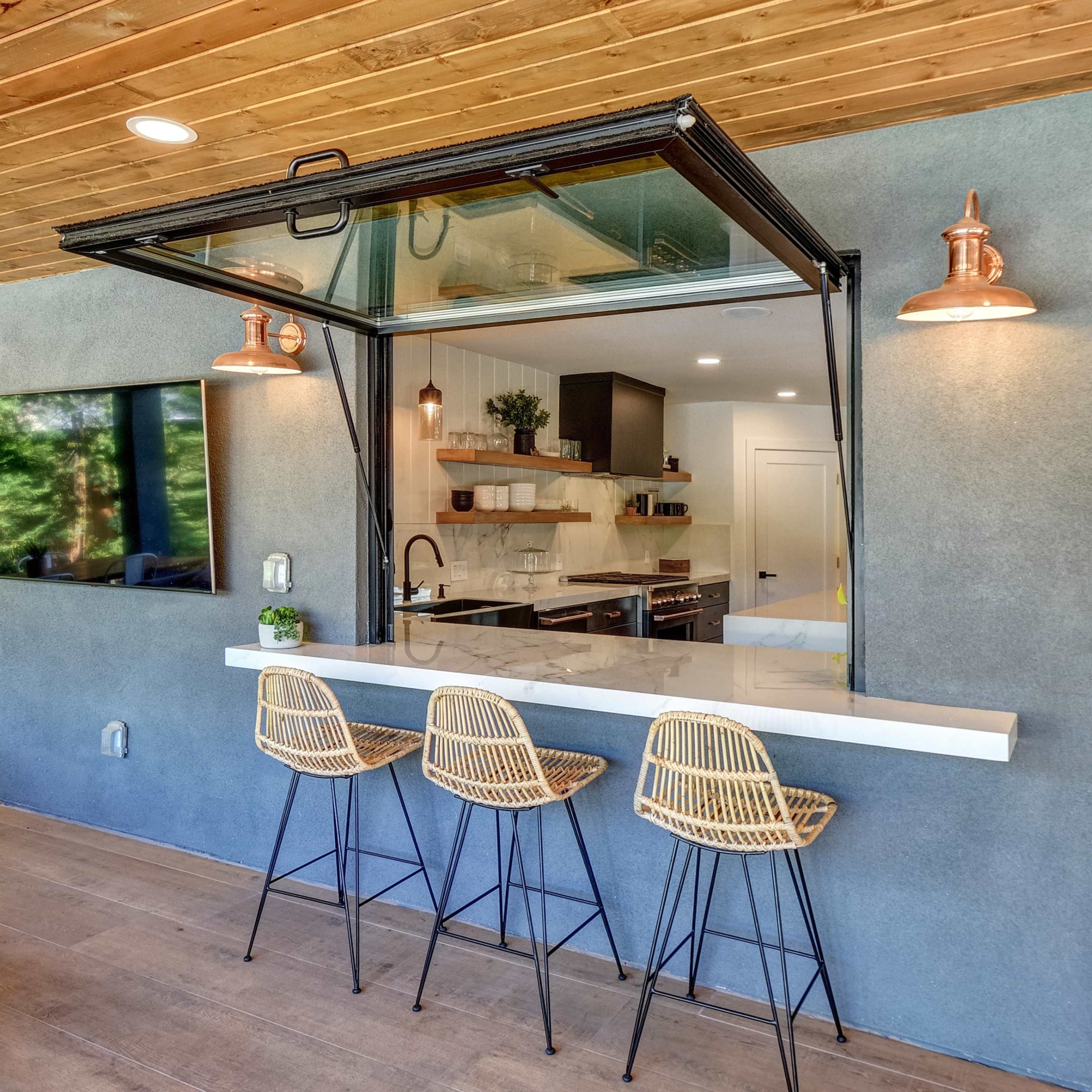 An open kitchen with a bar counter and three woven stools, featuring large glass windows that connect the indoor space to the outdoor area.
