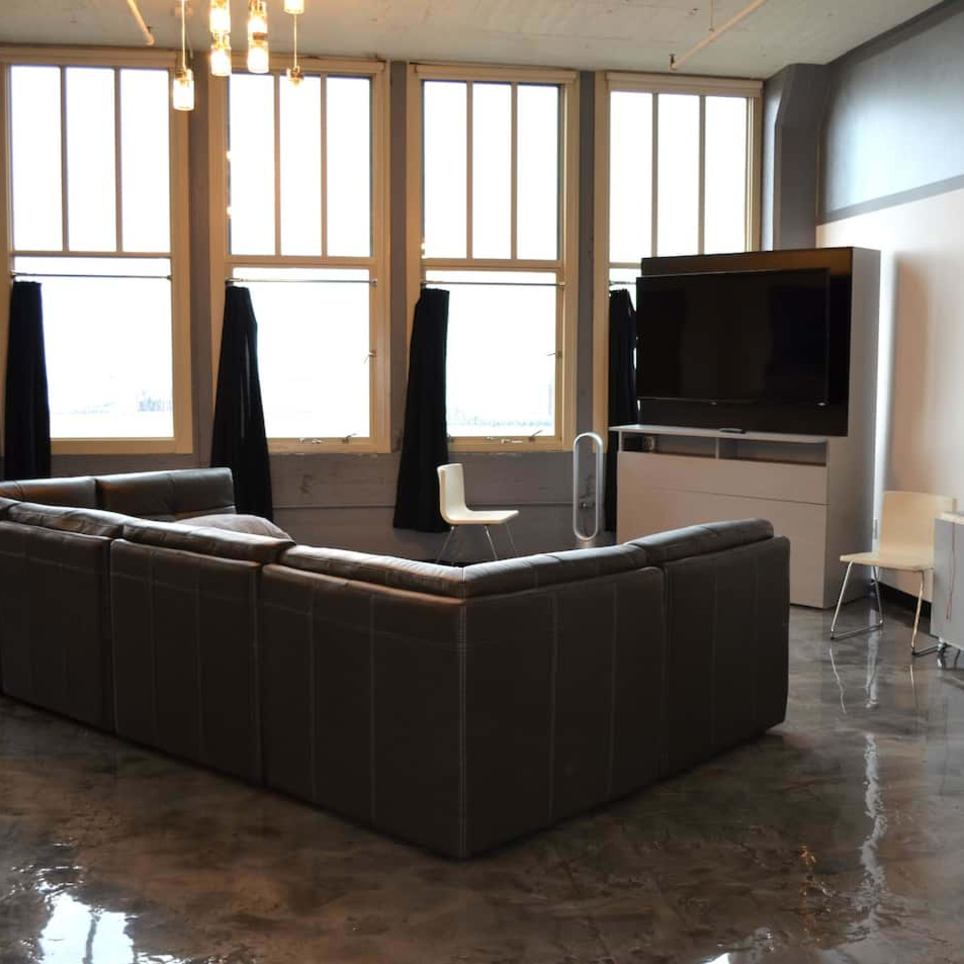 A modern living room featuring a large gray sectional sofa, a flat-screen TV mounted on the wall, and large windows that allow natural light to fill the space.