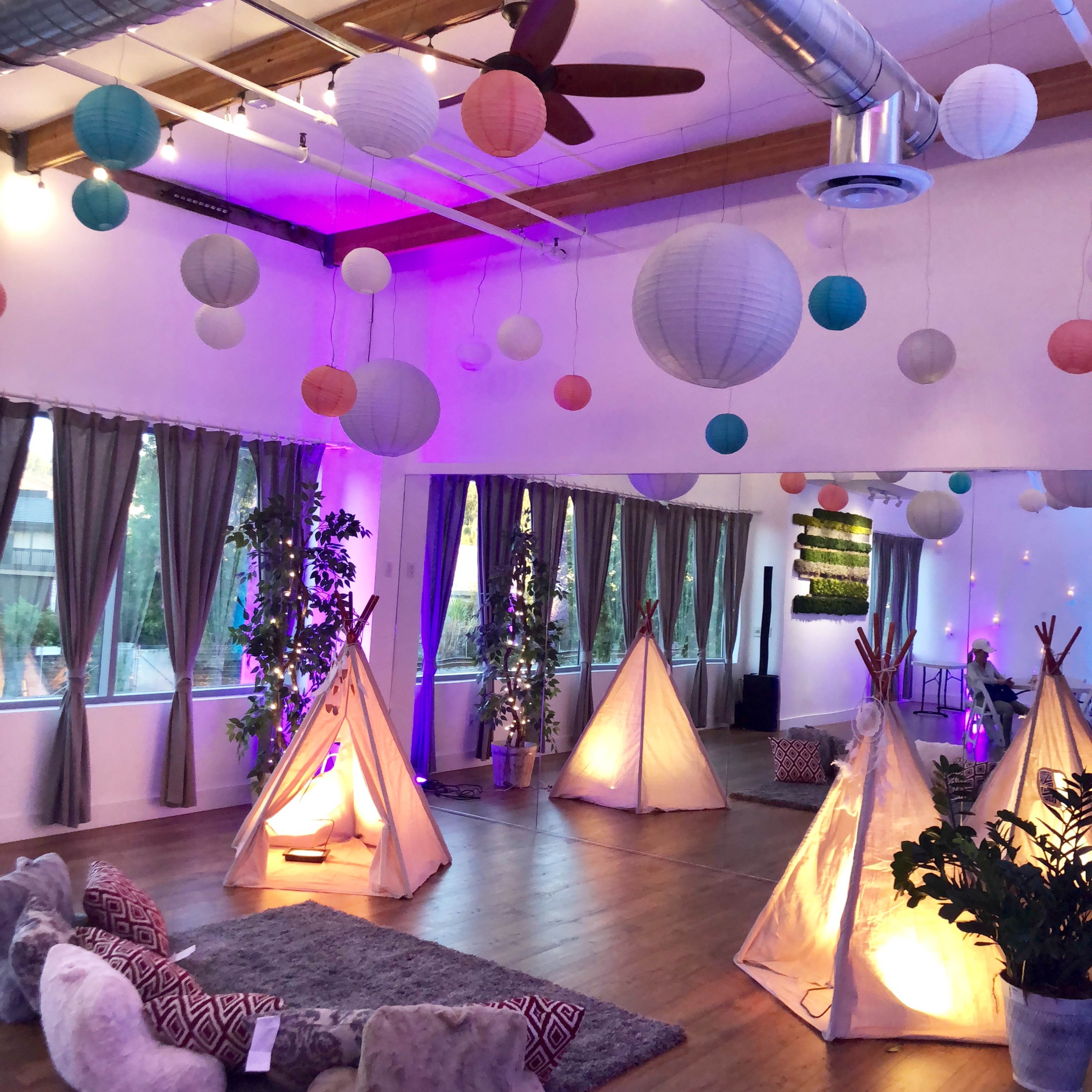 A spacious room is decorated with paper lanterns and features several illuminated teepees, with plush seating arranged on the floor.