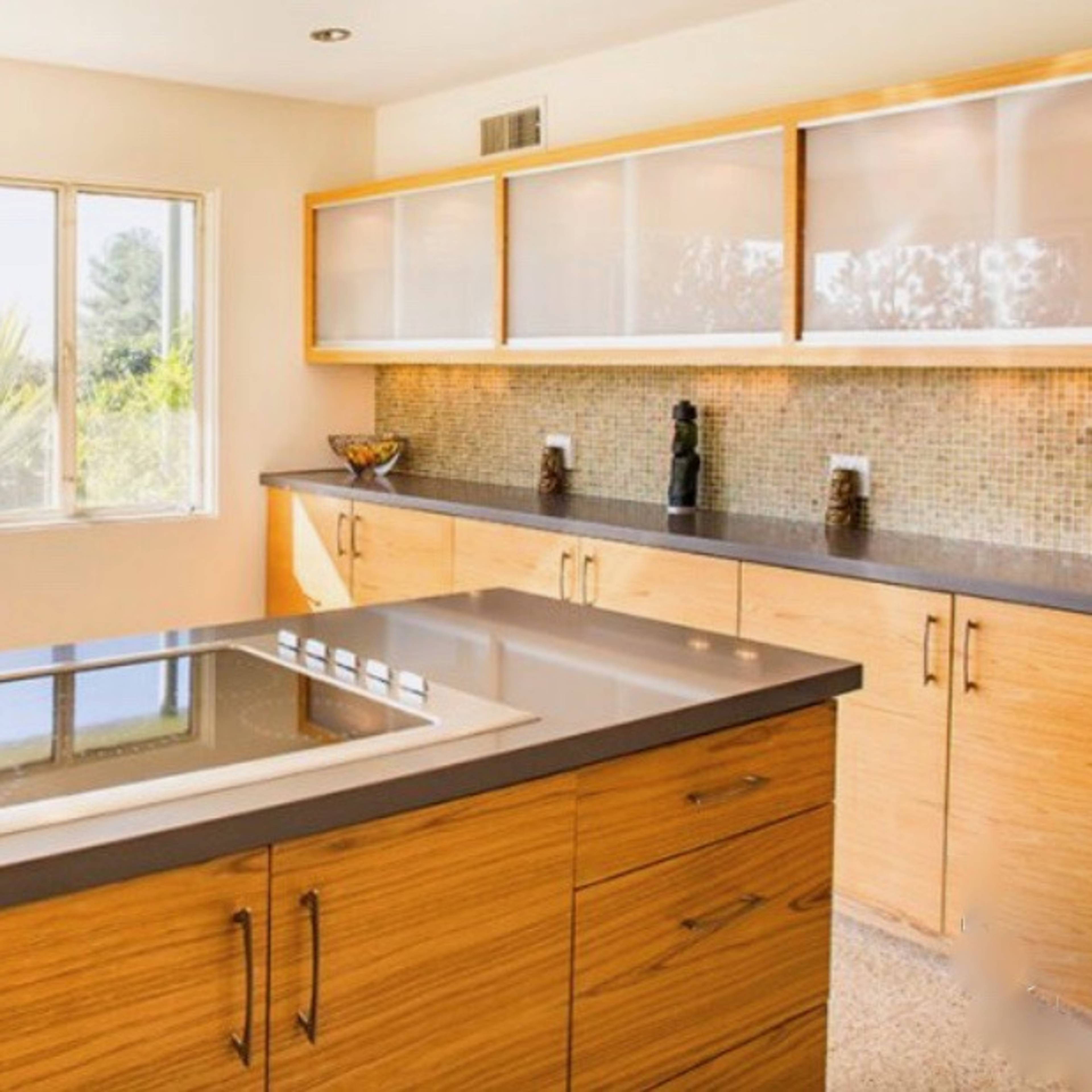 A modern kitchen with wooden cabinetry and a central island, overlooking a bright outdoor area with a dining table and palm trees visible through large windows.