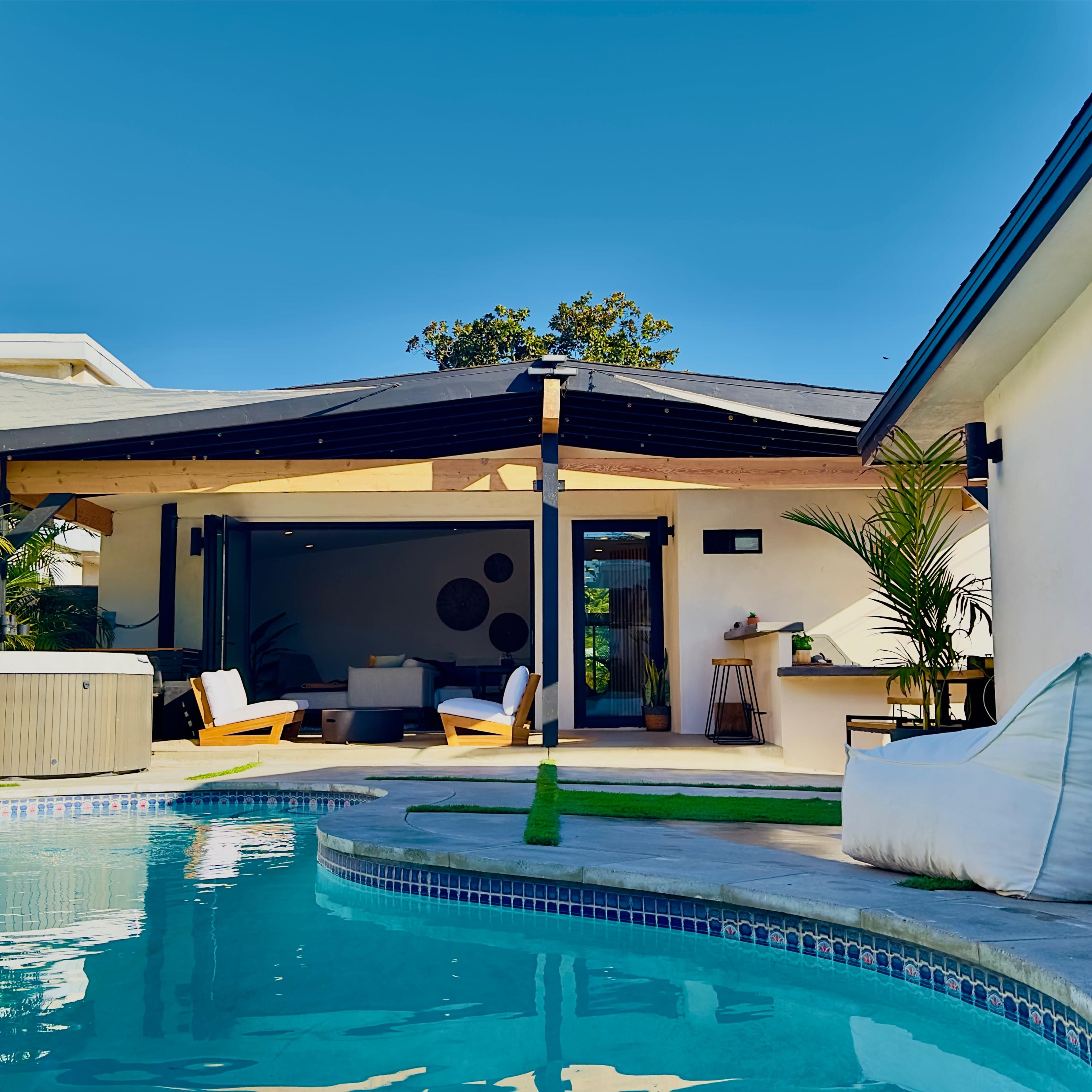 A modern outdoor space features a swimming pool, a covered patio with seating, and a hot tub surrounded by greenery.
