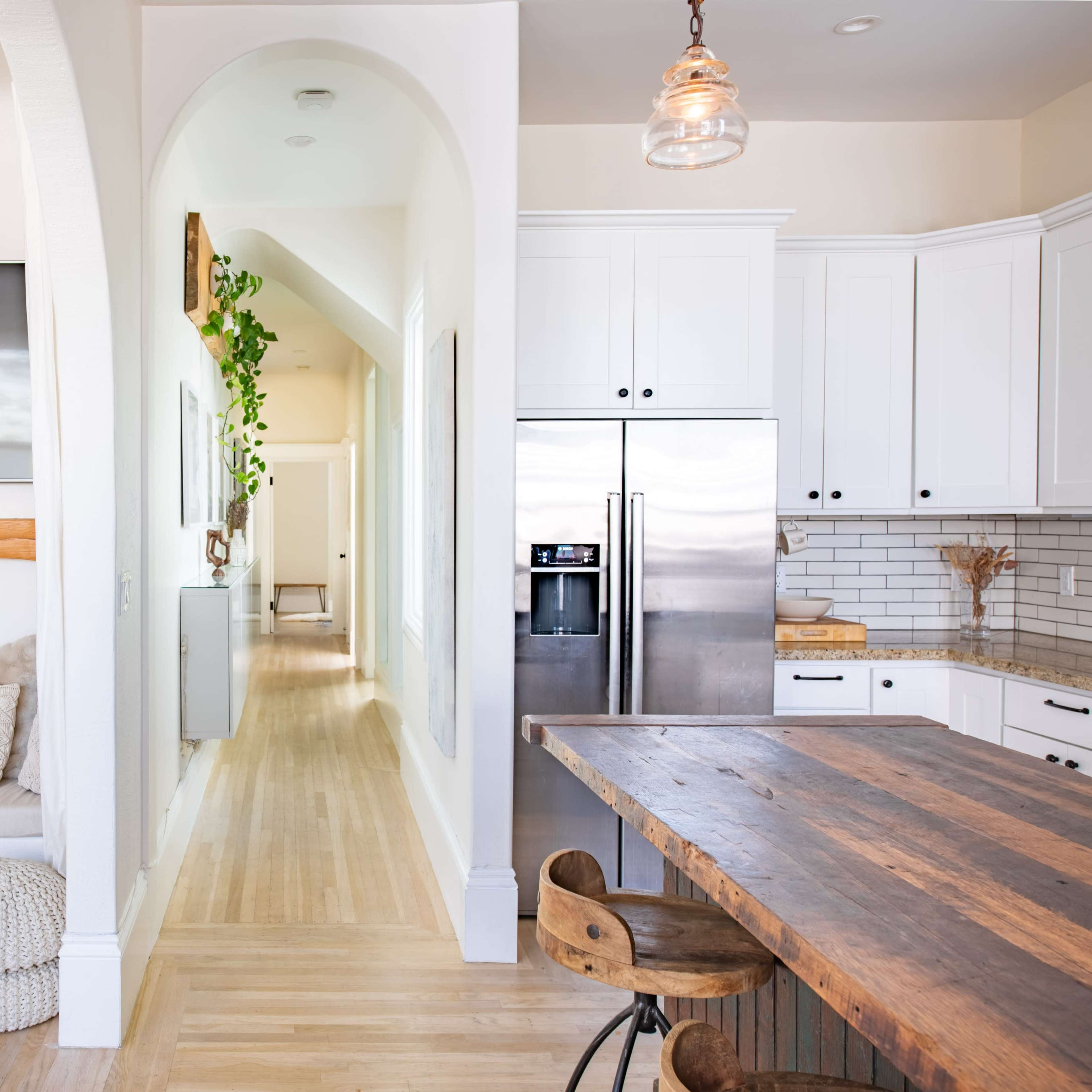 A modern living space with a cozy seating area beside a wall-mounted TV, an open kitchen featuring stainless steel appliances, and a light-filled hallway leading to another room.