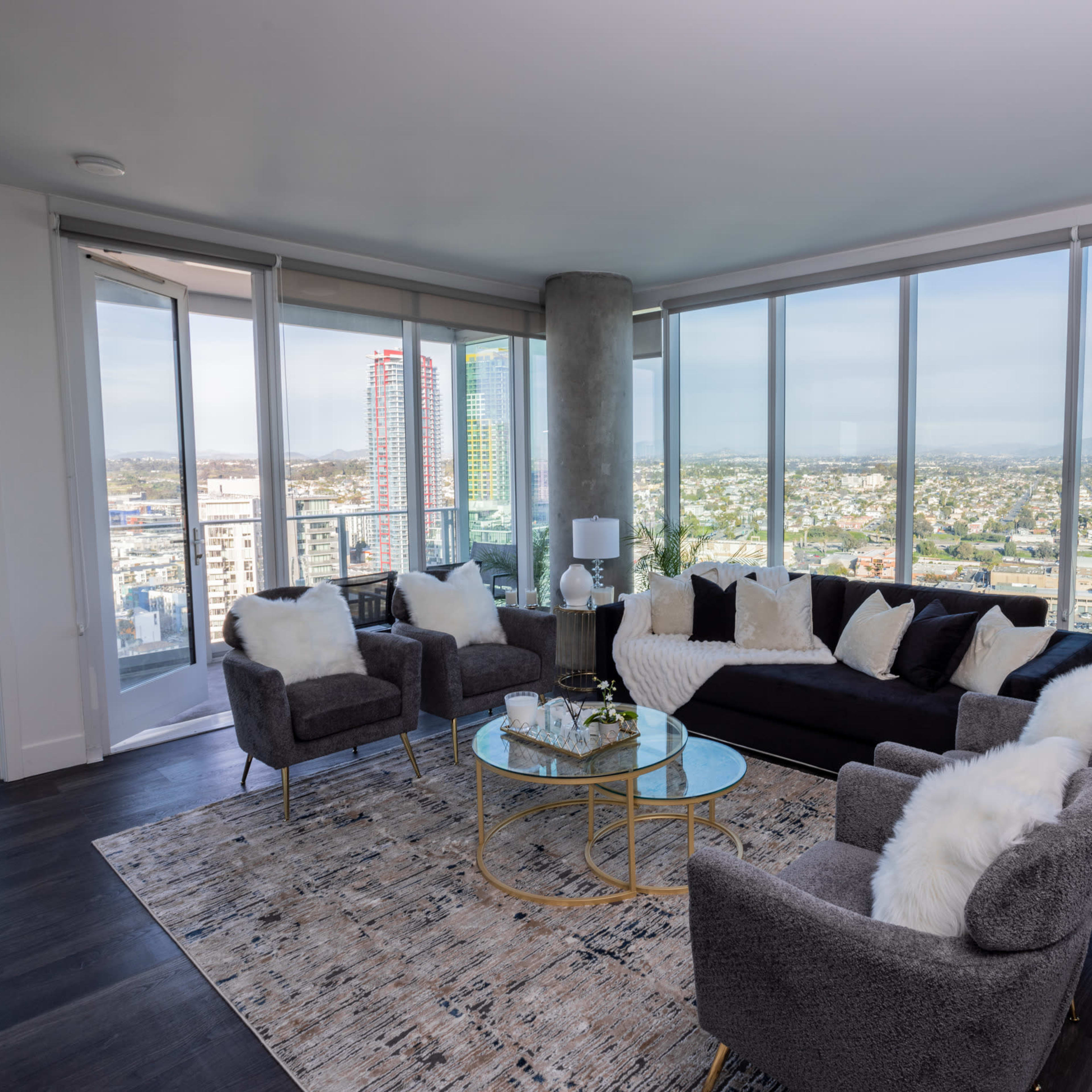 A modern living room with a gray sectional sofa and armchairs, a glass coffee table, and large windows offering a panoramic view of a city skyline.