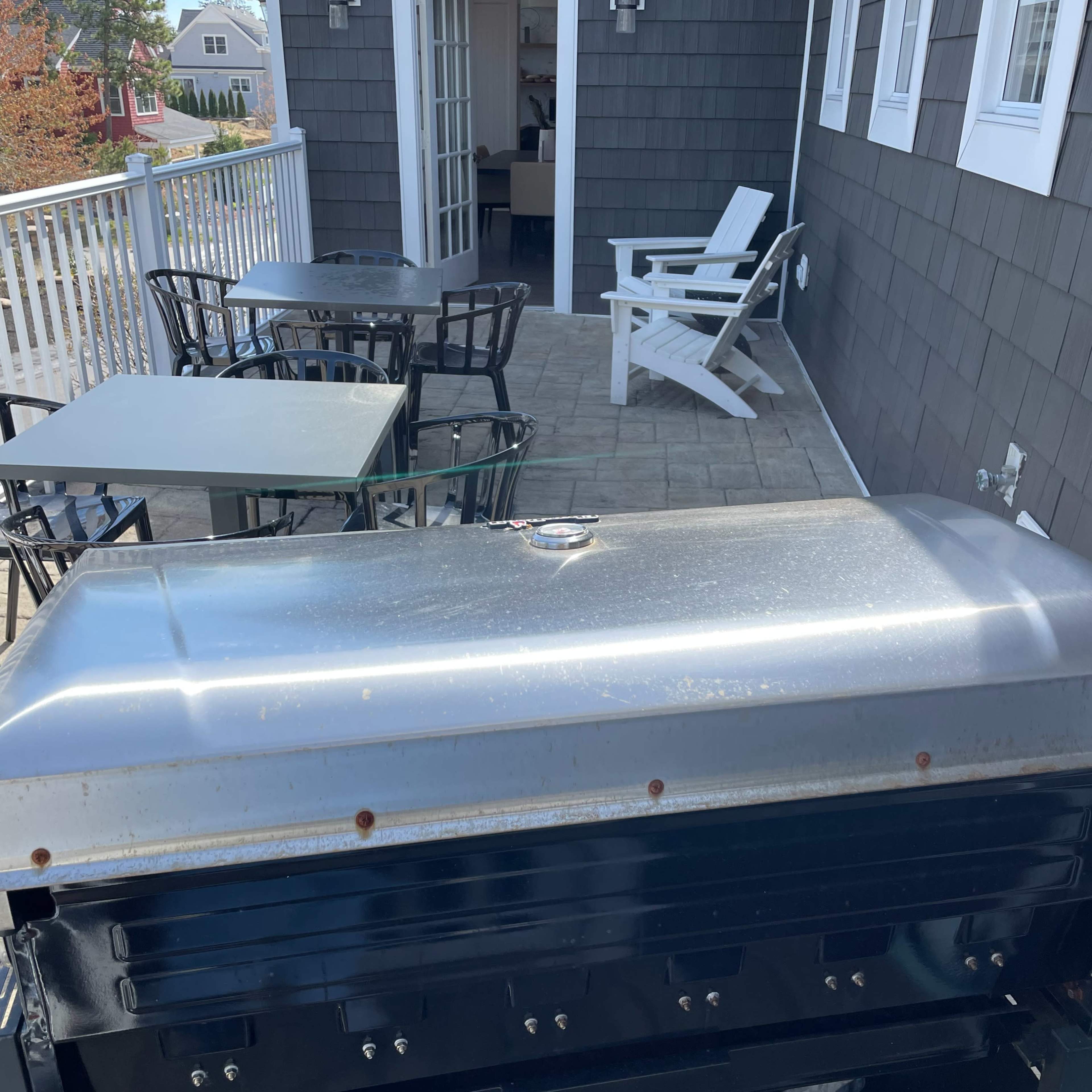 A stainless steel grill is positioned on a balcony overlooking a table and chairs set against a backdrop of a house with gray siding and a door leading inside.