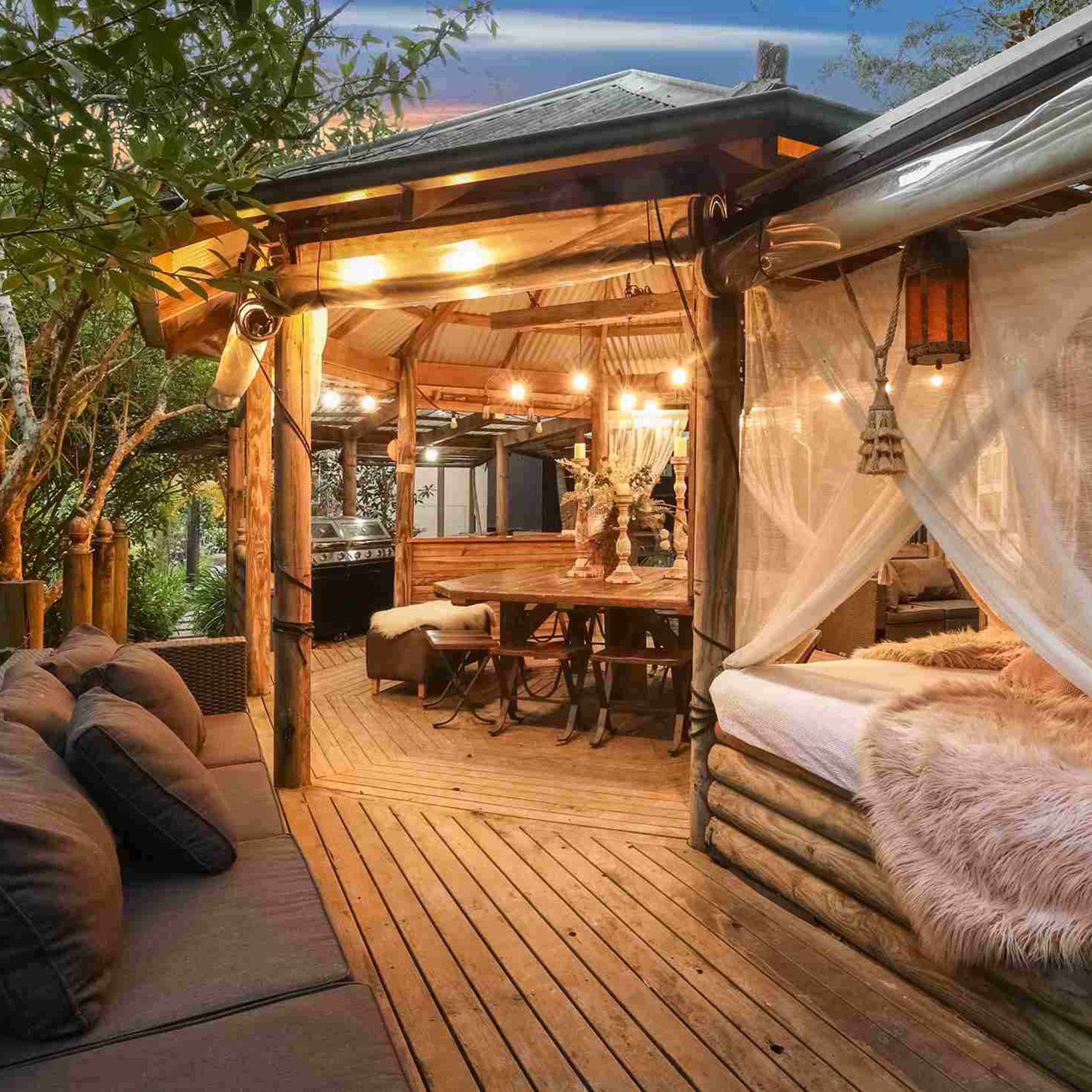 A wooden deck with a cozy seating area, a dining space, and a bed framed by sheer curtains, surrounded by greenery and softly glowing lights.