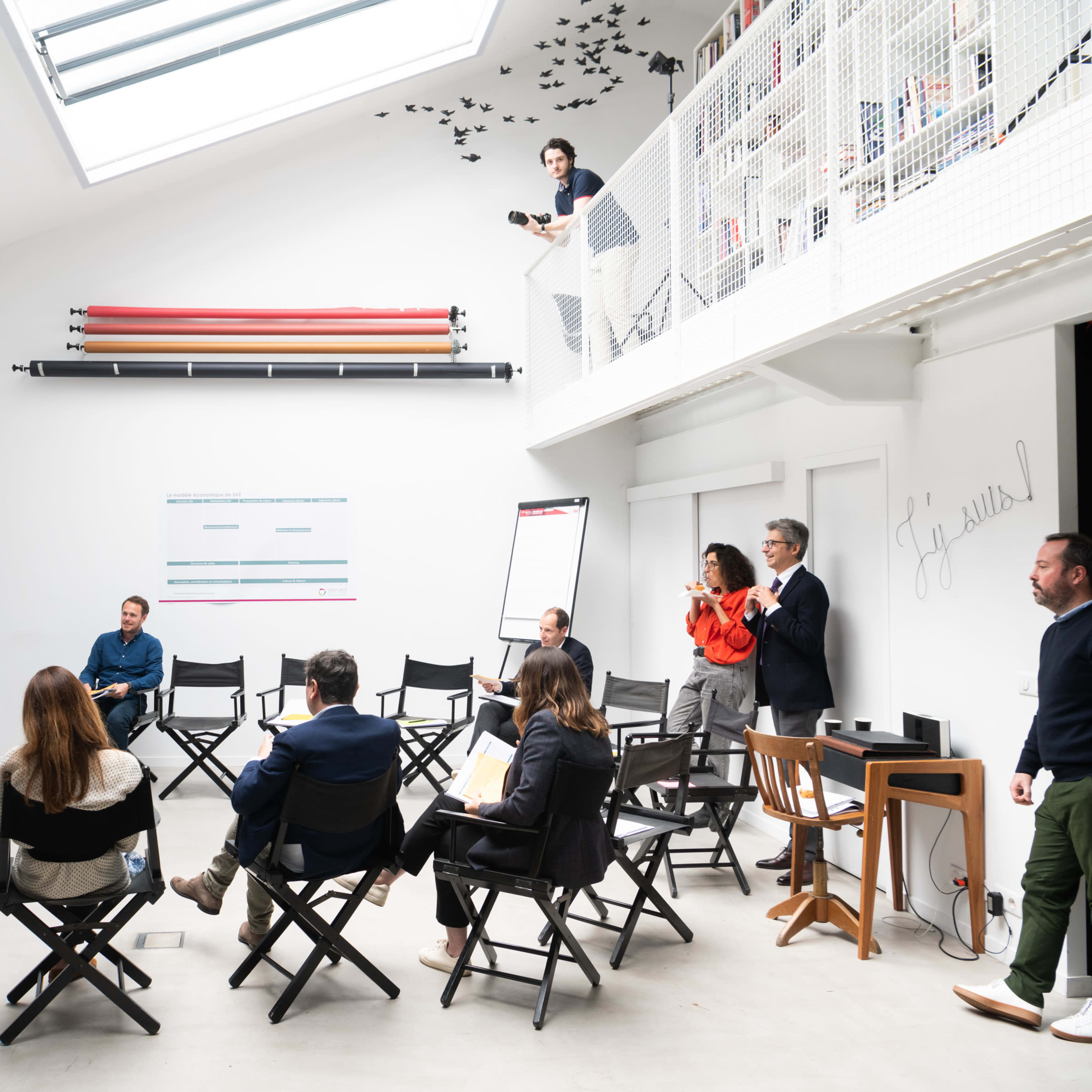 A group of people engage in a meeting in a bright, modern room with a skylight and a whiteboard.