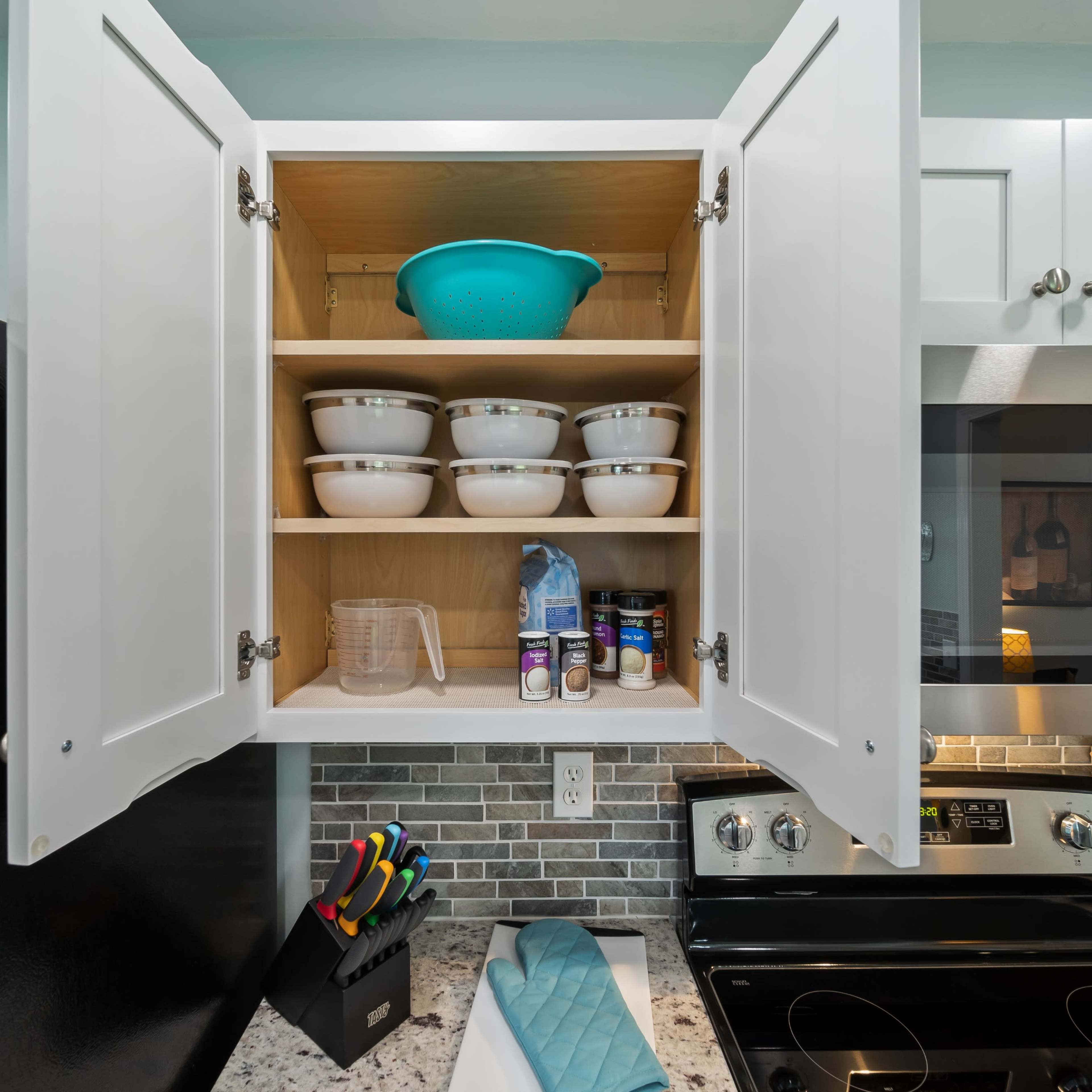 A kitchen cabinet is open, displaying bowls, a measuring cup, and spice jars arranged neatly on shelves above a stovetop and a countertop with cooking utensils.