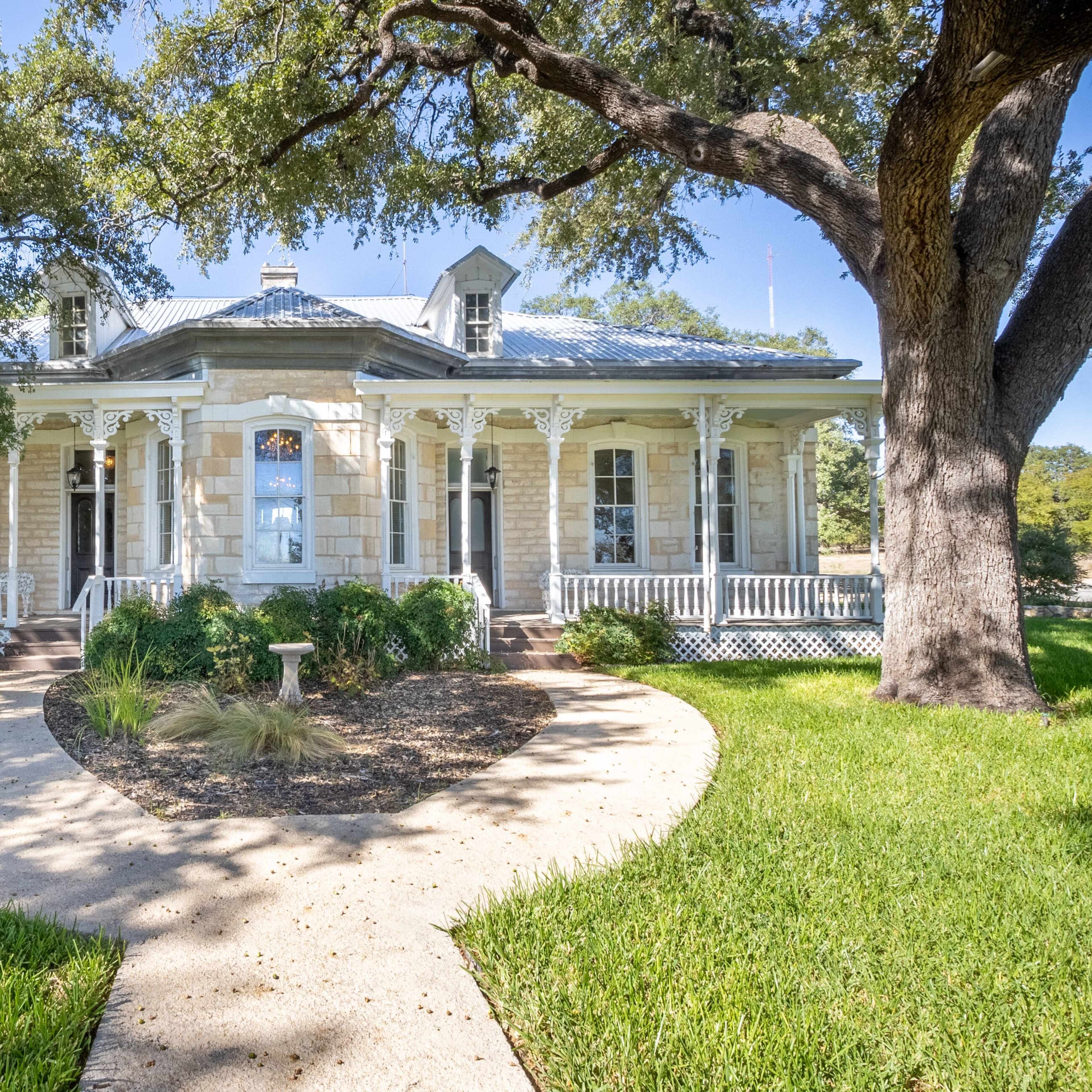 A historic stone house with a wraparound porch, surrounded by green grass and mature trees.
