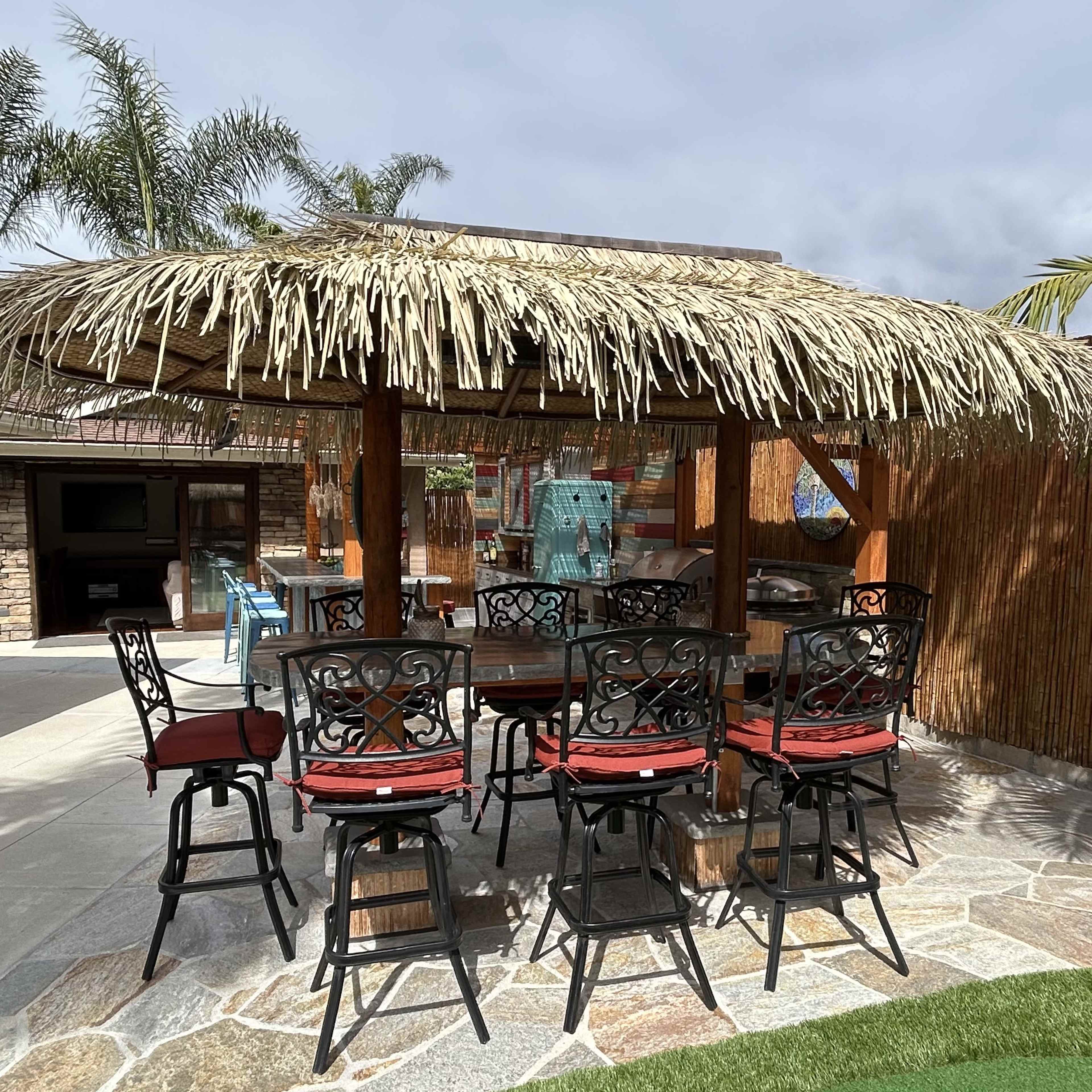 A thatched-roof gazebo with a wooden table and chairs is surrounded by tropical landscaping and stone flooring.