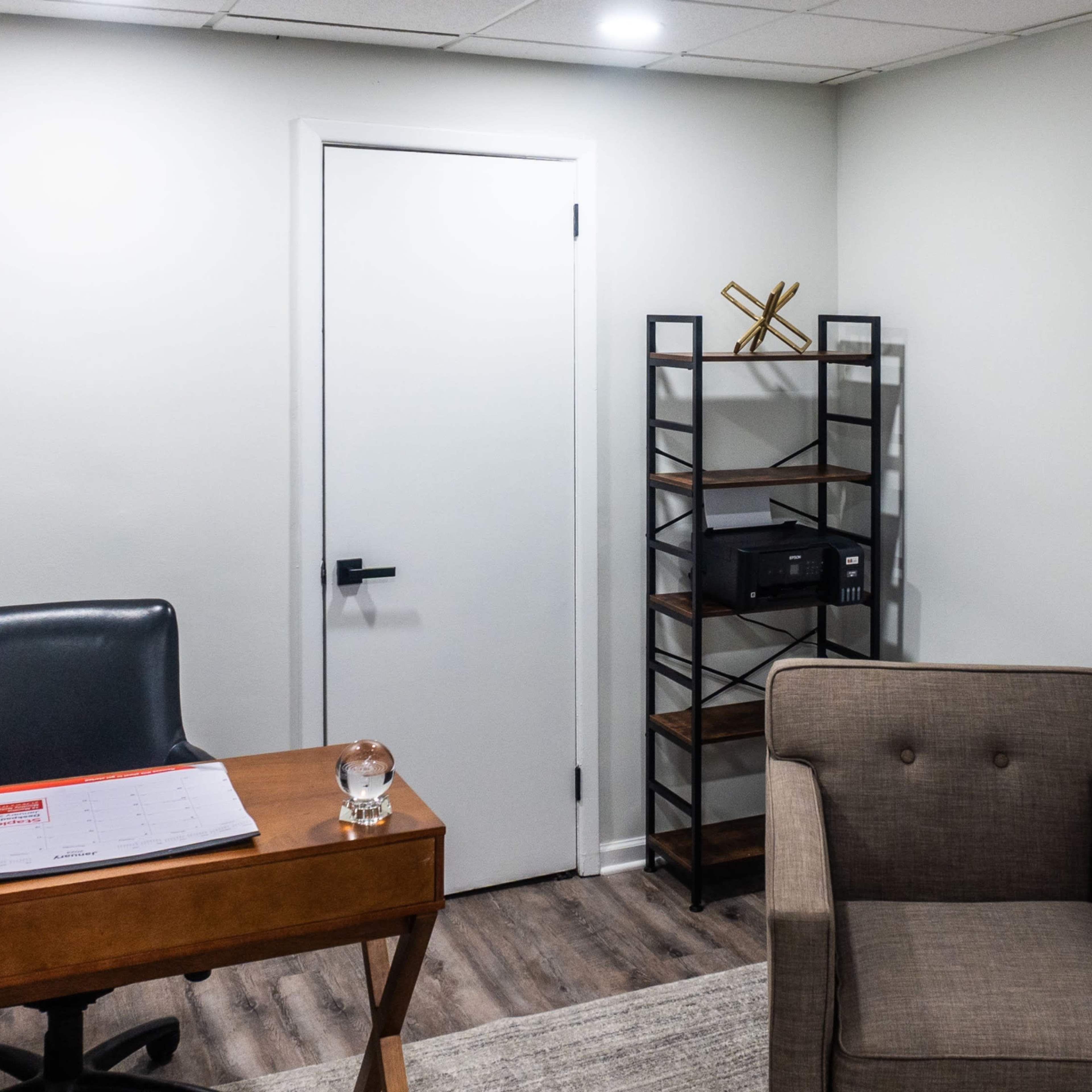 An office space features a wooden desk with a calendar, a black leather chair, a gray armchair, and a metal shelving unit against a light-colored wall.