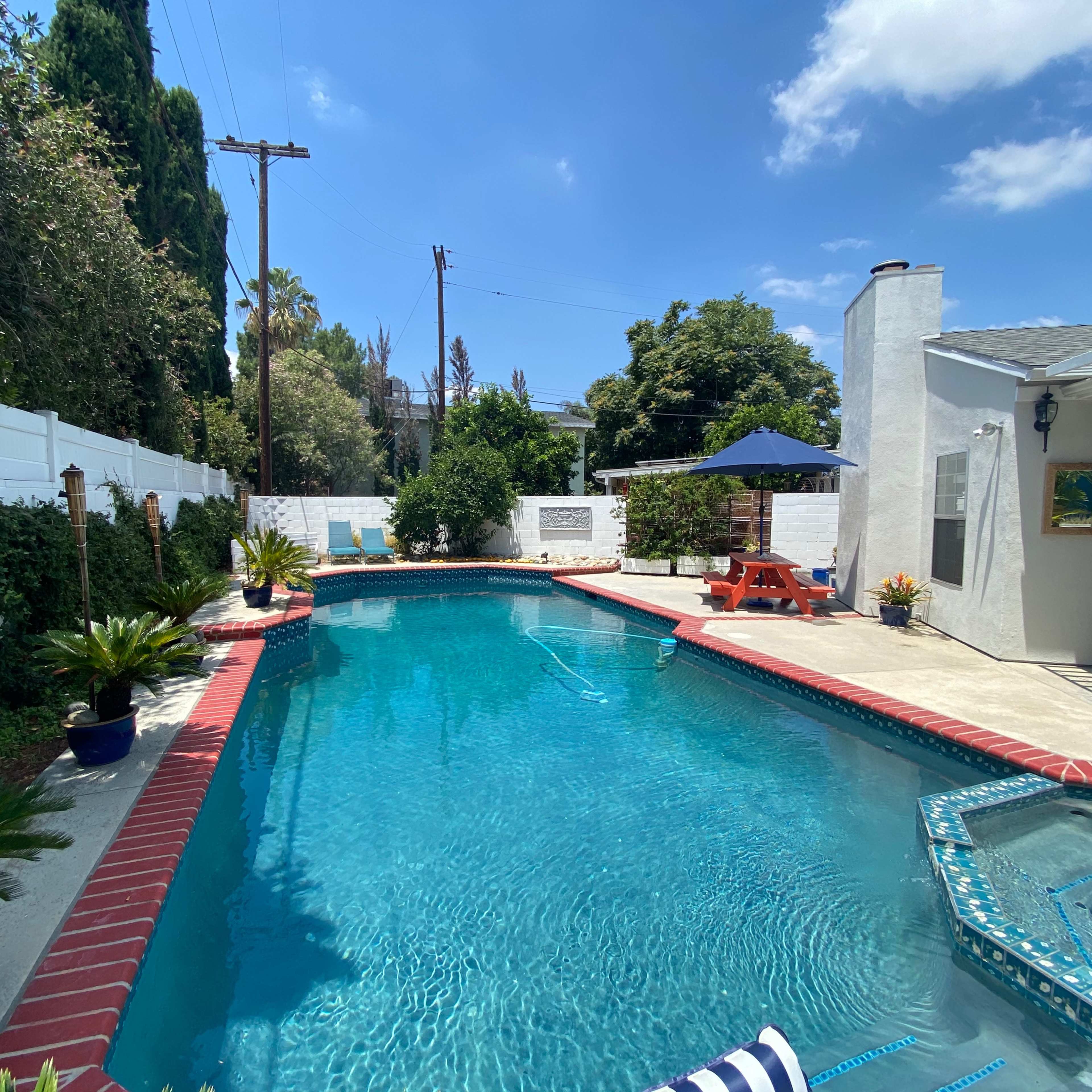 A backyard pool area surrounded by greenery, featuring a clear blue pool with a Jacuzzi, loungers, and a shaded seating area under an umbrella.