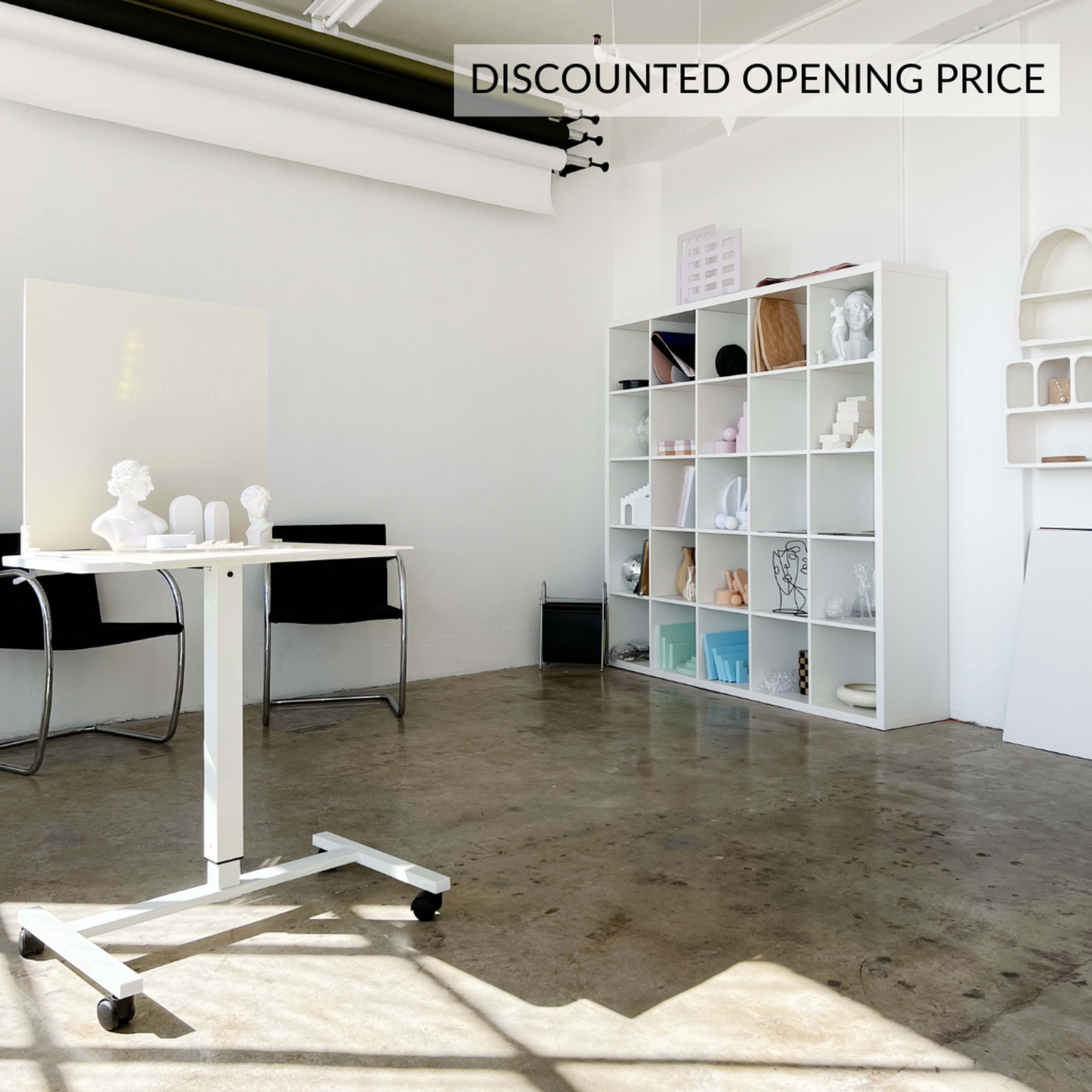 A modern, minimalist studio space with a white table on wheels, black chairs, and a white shelving unit filled with various decorative objects.
