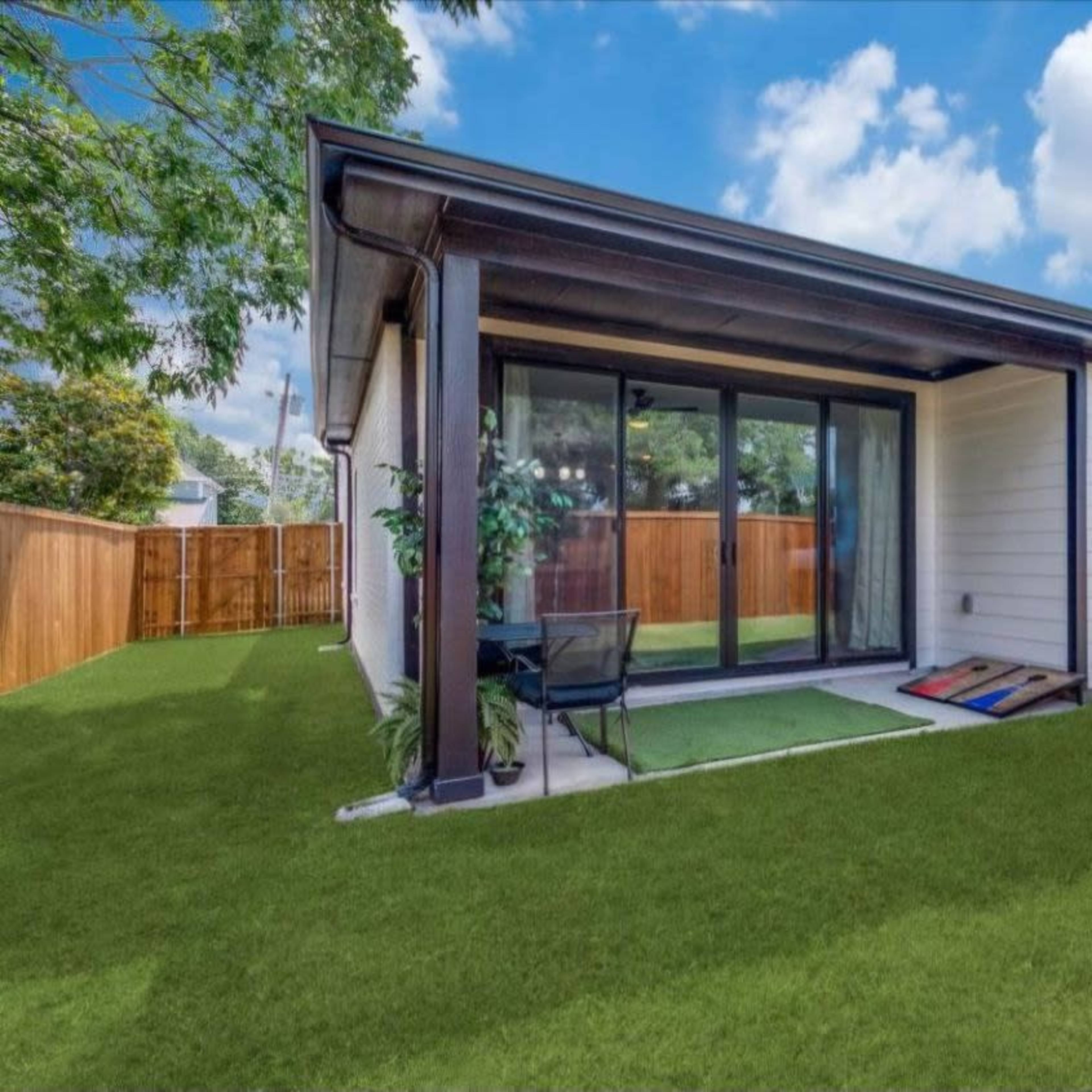 A small modern house features a patio area with sliding glass doors opening to a grassy backyard surrounded by a wooden fence.