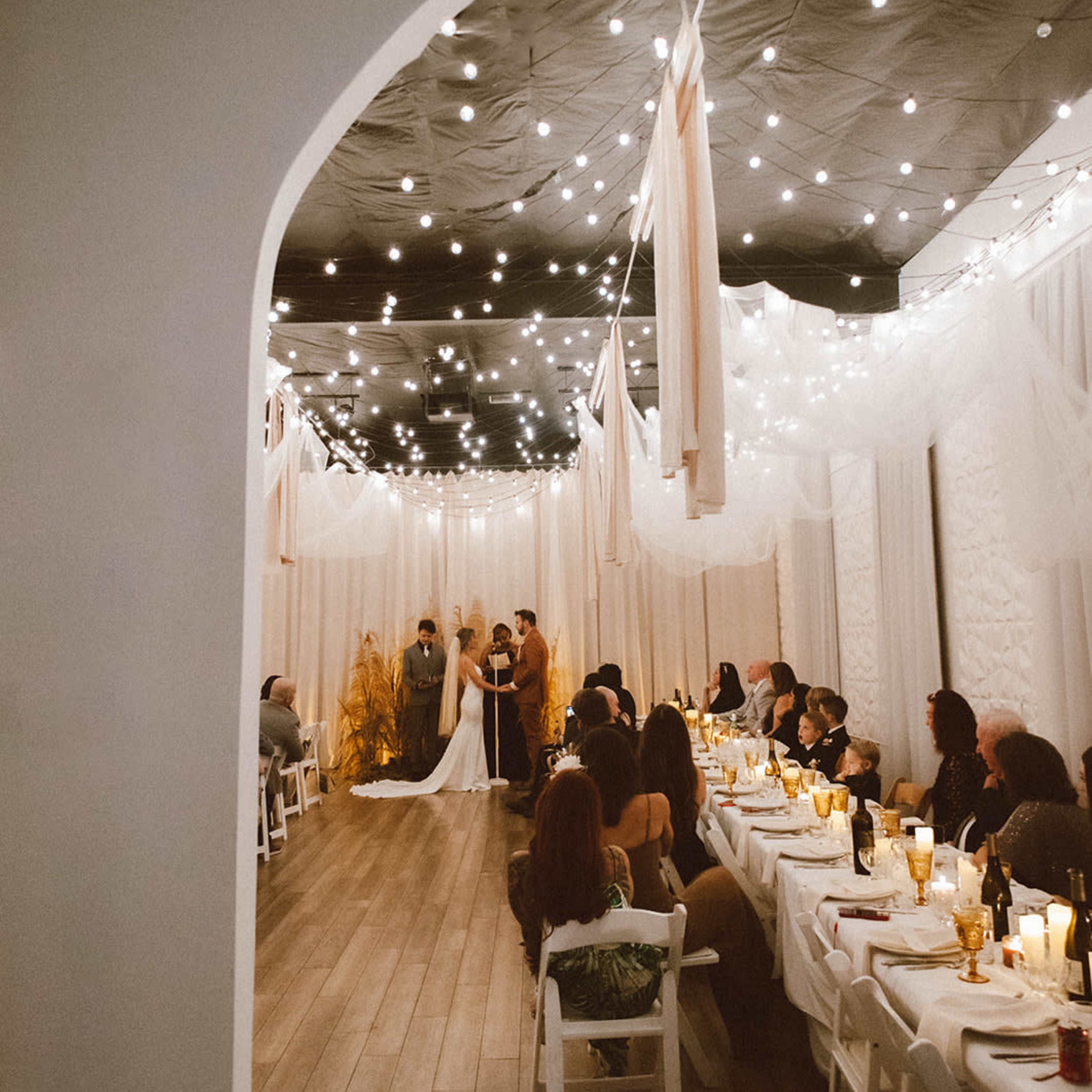 A wedding ceremony takes place under string lights in a decorated indoor space with guests seated at long tables.
