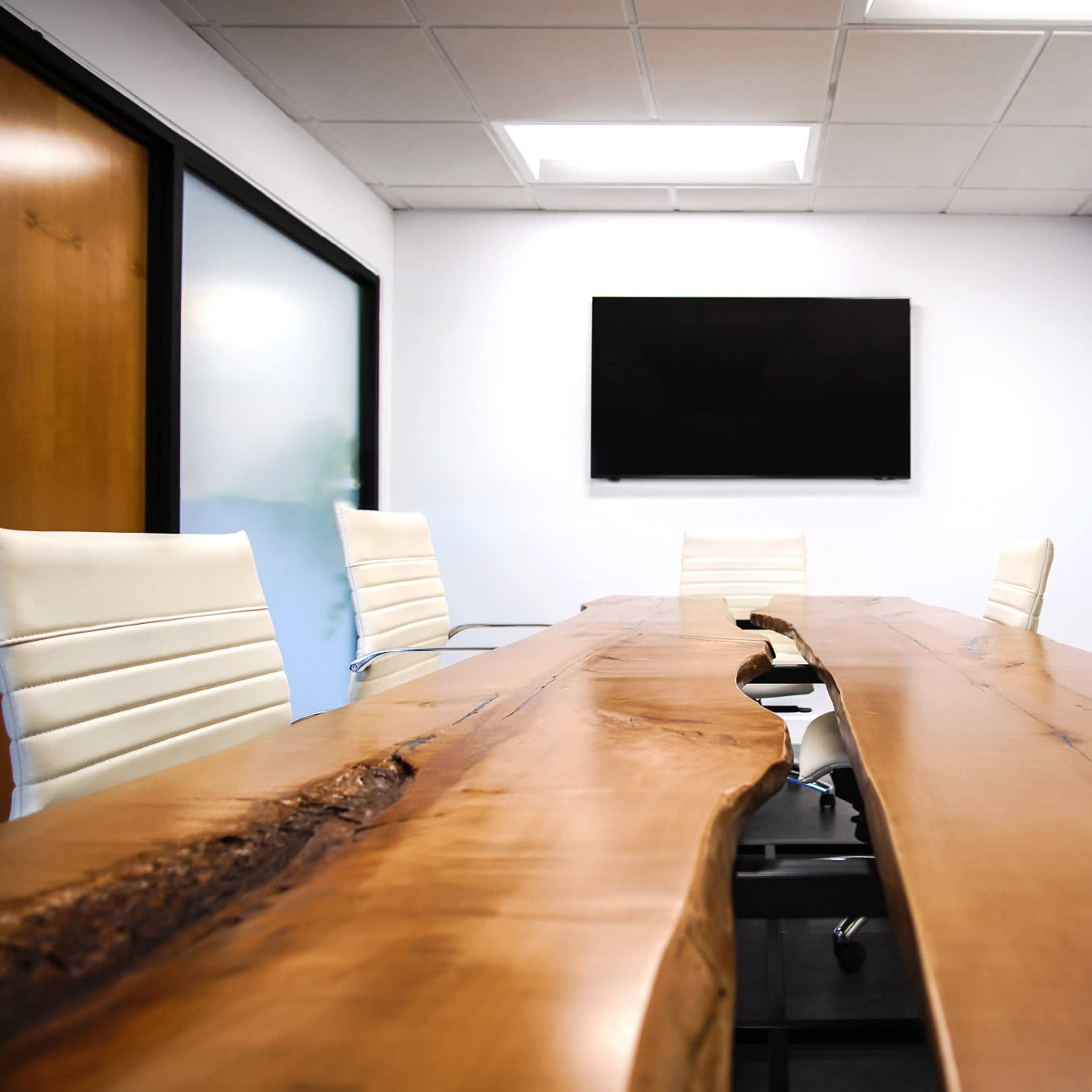 A long wooden conference table is set with white chairs in a modern meeting room featuring a wall-mounted television and colorful geometric wall decor.