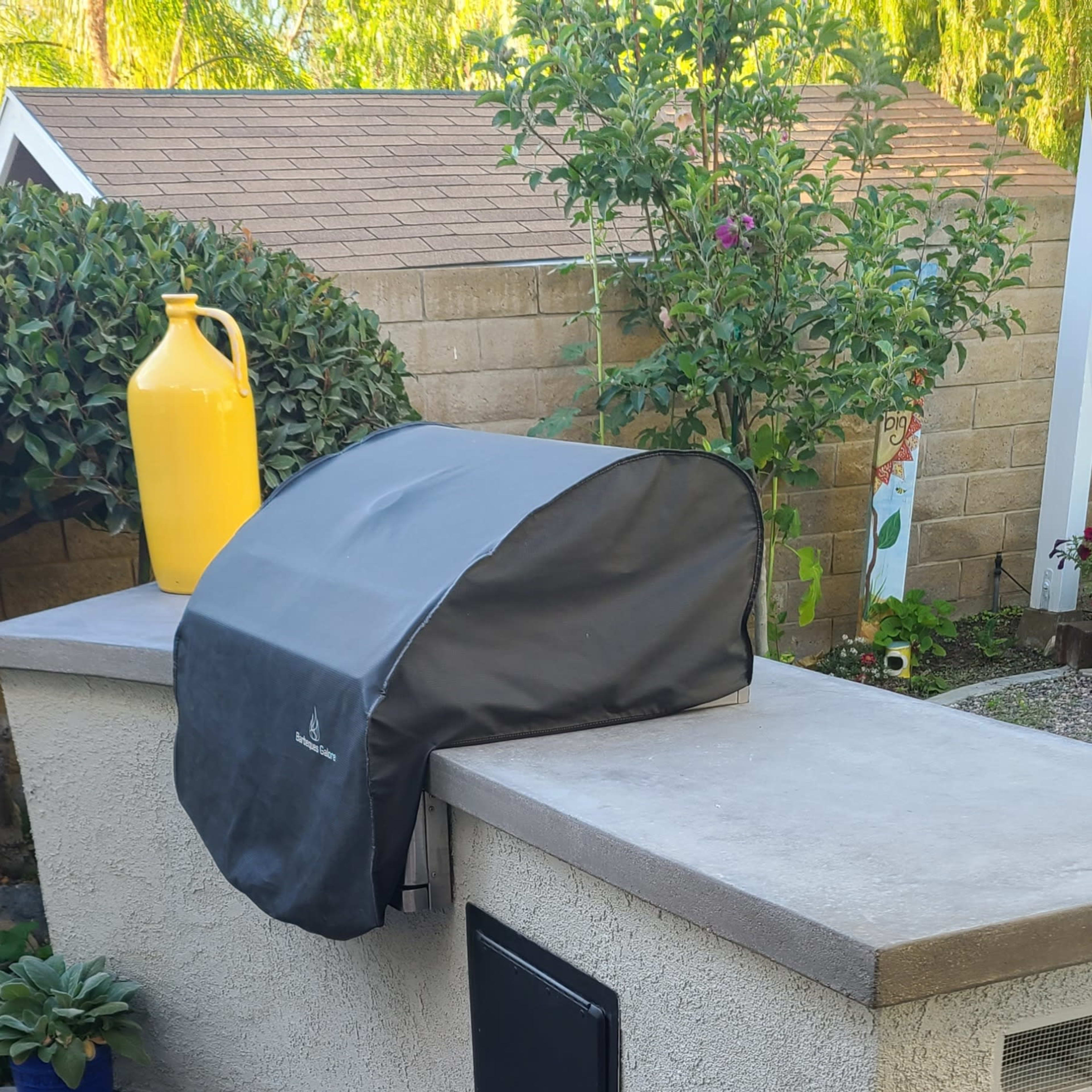A grill is covered with a black cover on a concrete outdoor countertop next to a yellow container and plants.