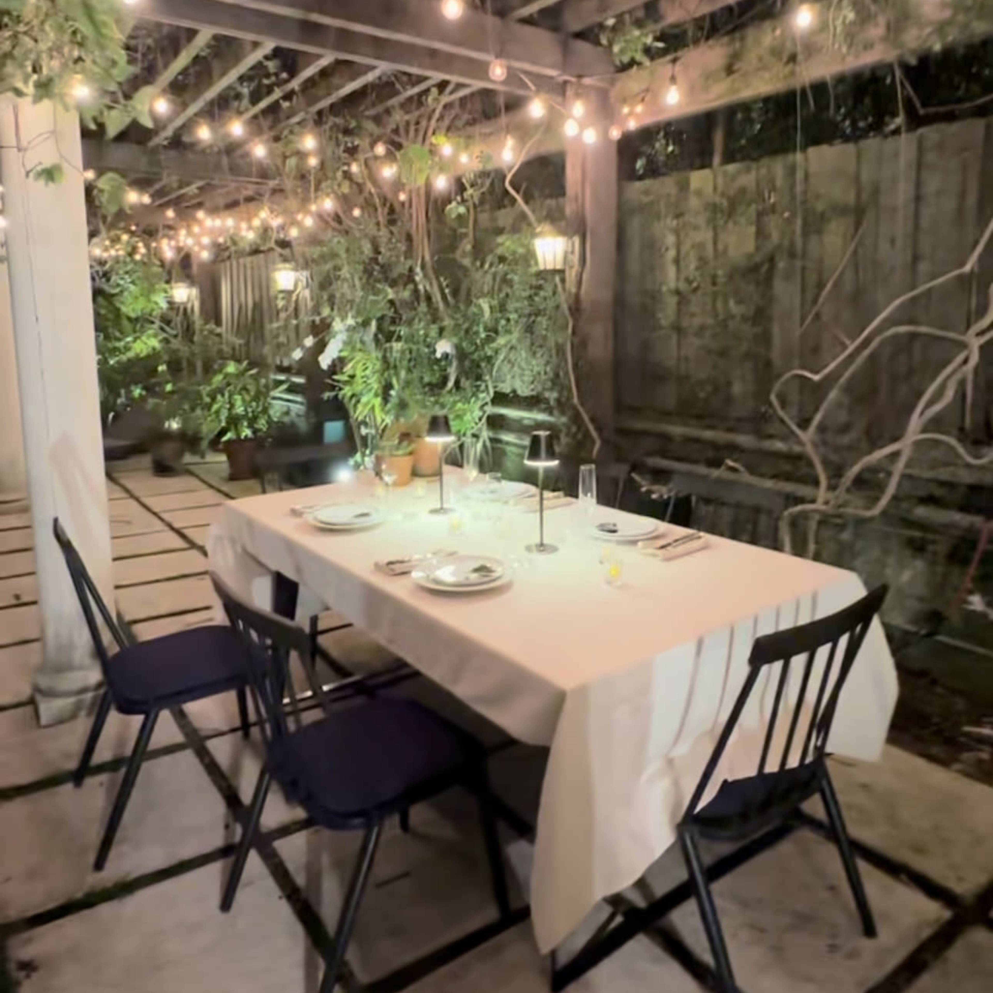 A table is set for dinner under a pergola adorned with string lights and surrounded by greenery.