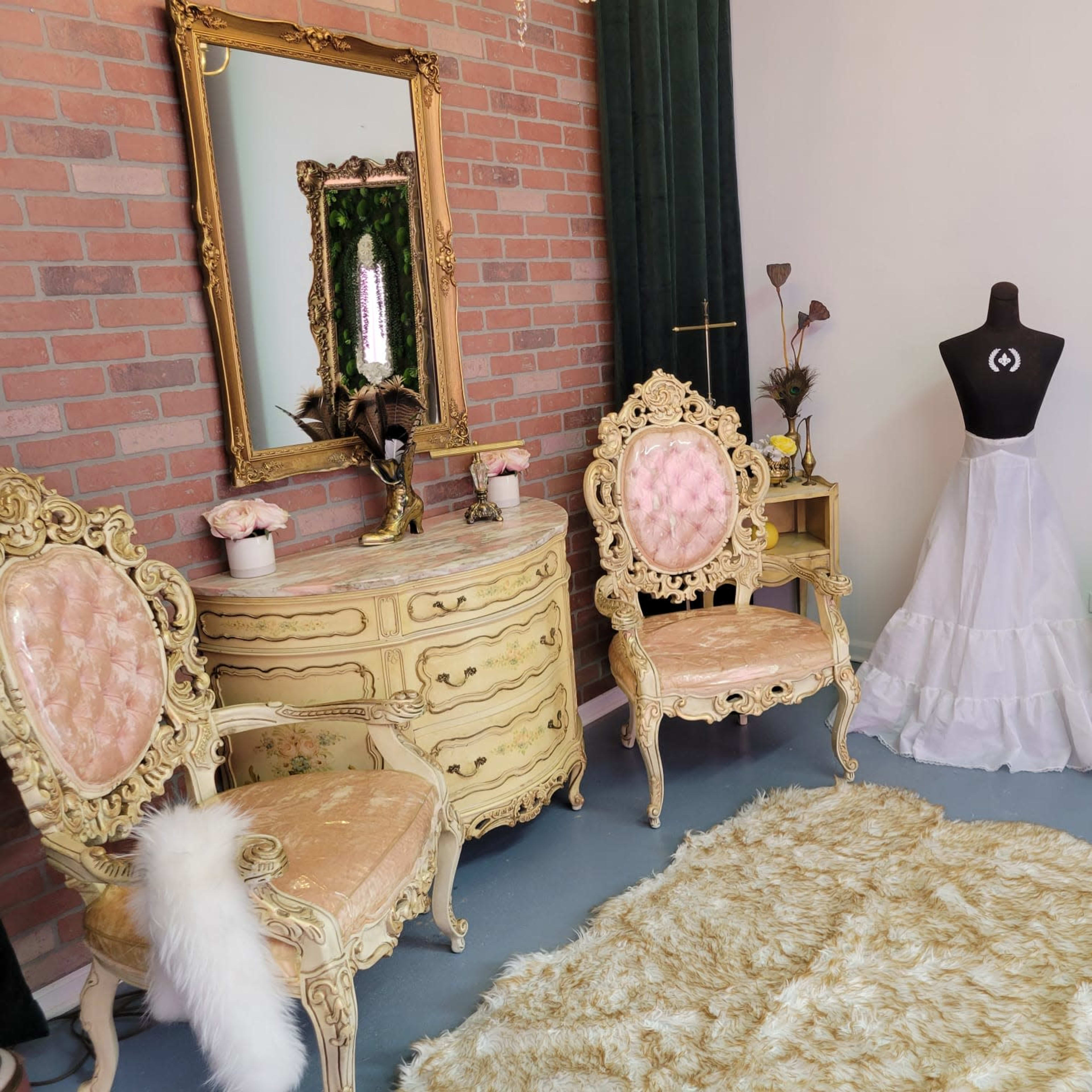 The interior features a vintage-style room with ornate golden furniture, including two chairs and a dresser, alongside a dress form and a decorative mirror surrounded by greenery.