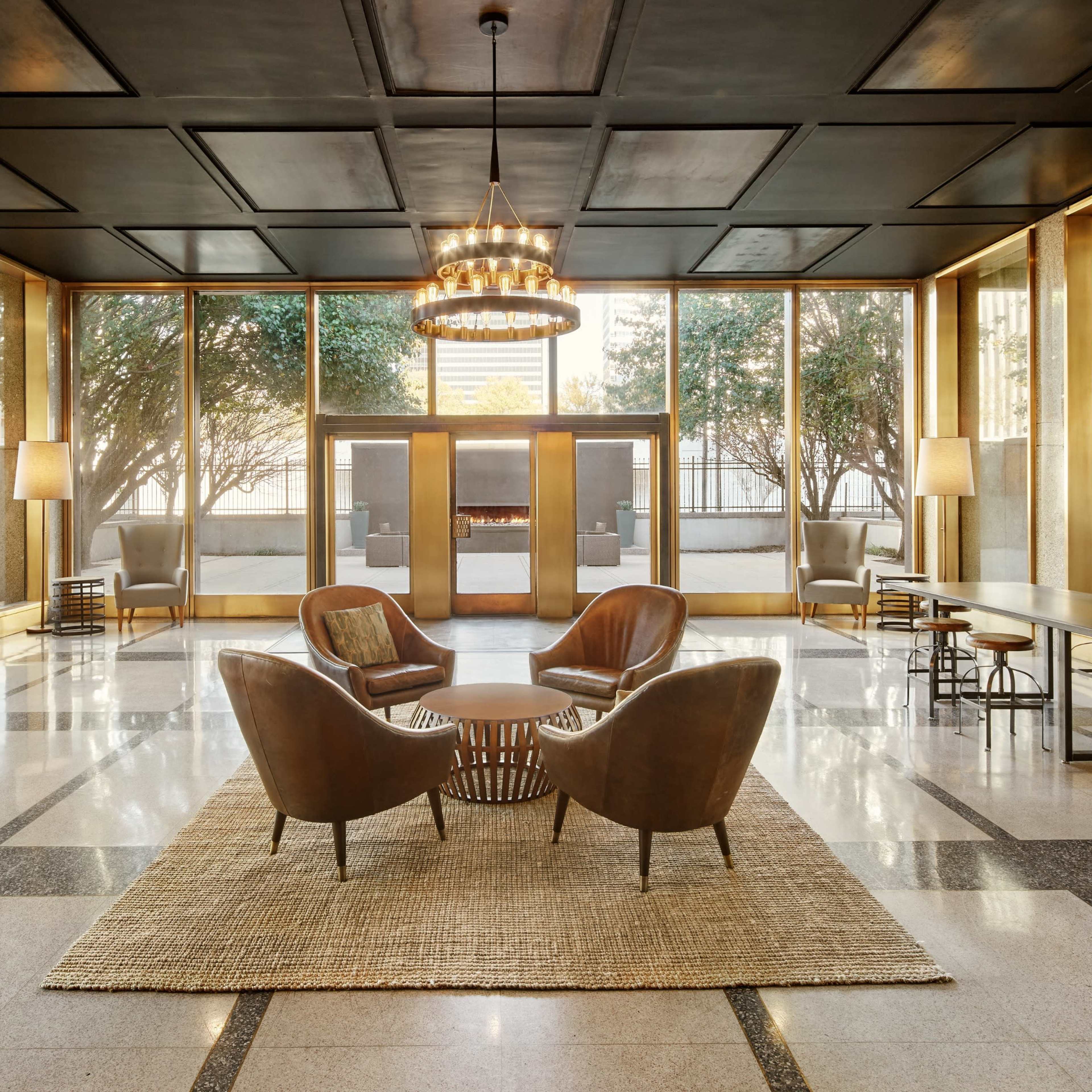 A modern lobby features two leather armchairs facing a round coffee table on a woven rug, with large windows providing an outside view and a chandelier hanging overhead.