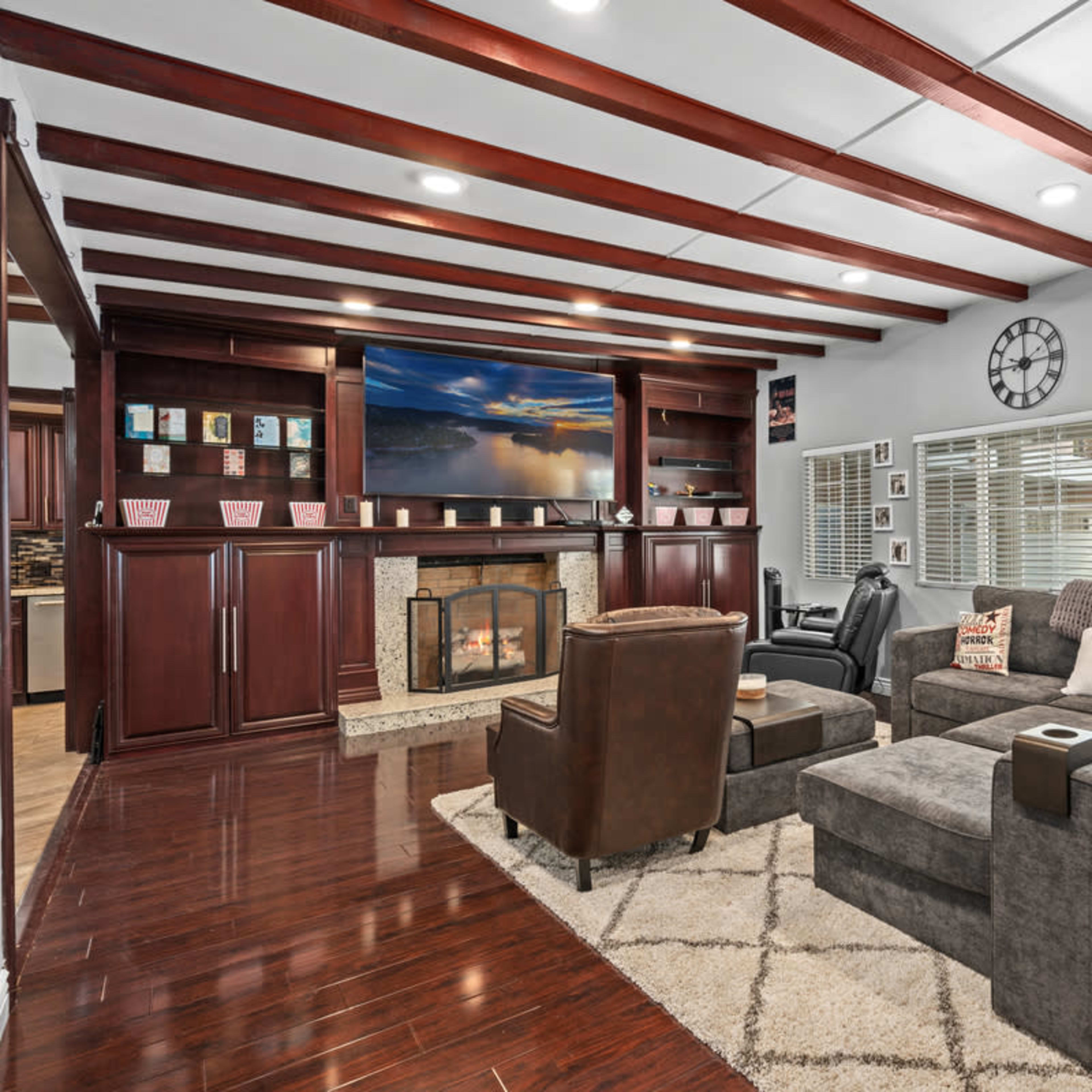 The living room features a sectional sofa, two armchairs, a wall-mounted TV above a fireplace, and wood beams on the ceiling, with large windows providing natural light.