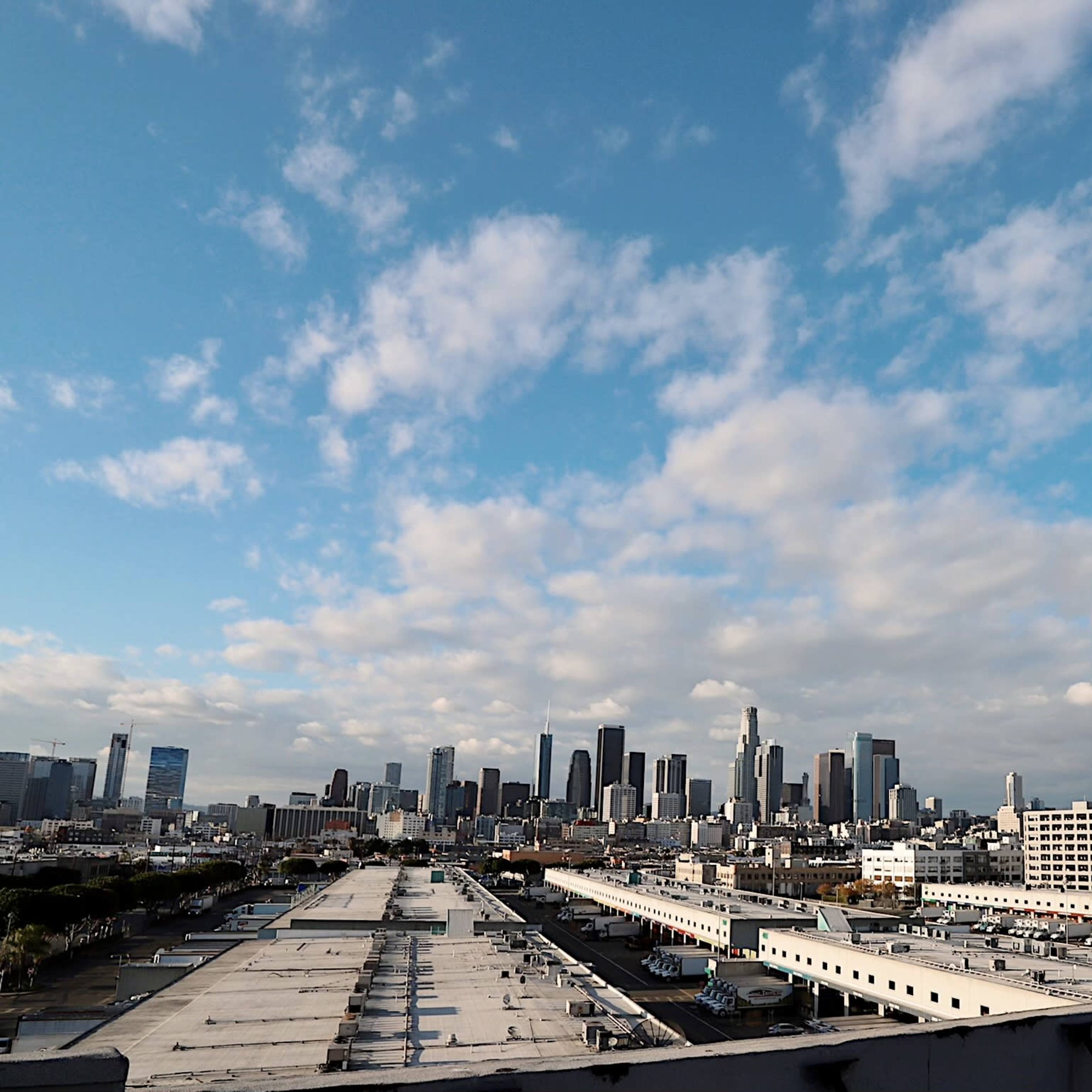A panoramic view of the Los Angeles skyline under a partly cloudy sky, with urban buildings and rooftops in the foreground.