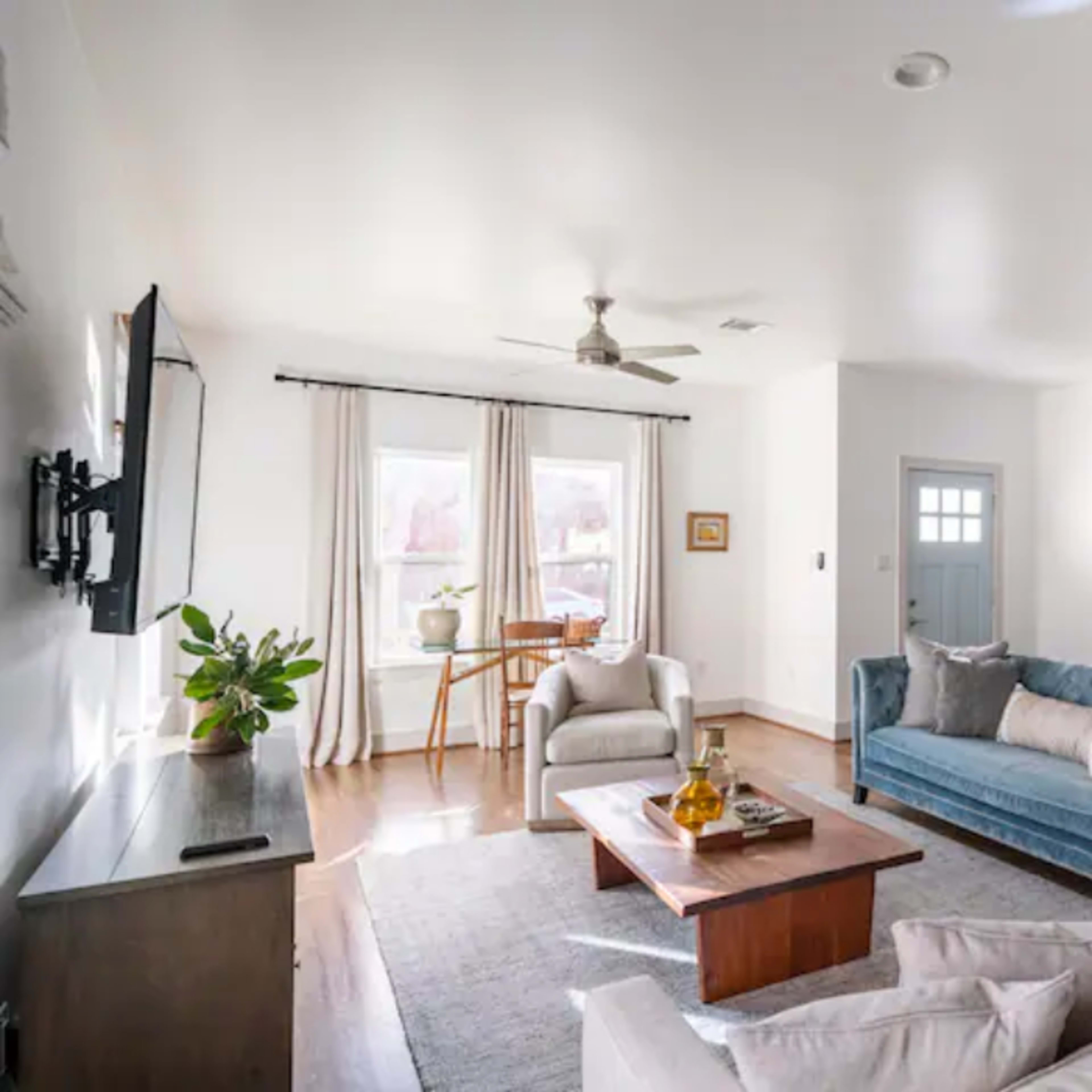 The living room features a blue velvet couch, a light-colored sofa, a coffee table, and a window with sheer curtains, complemented by a small dining table and light wood flooring.