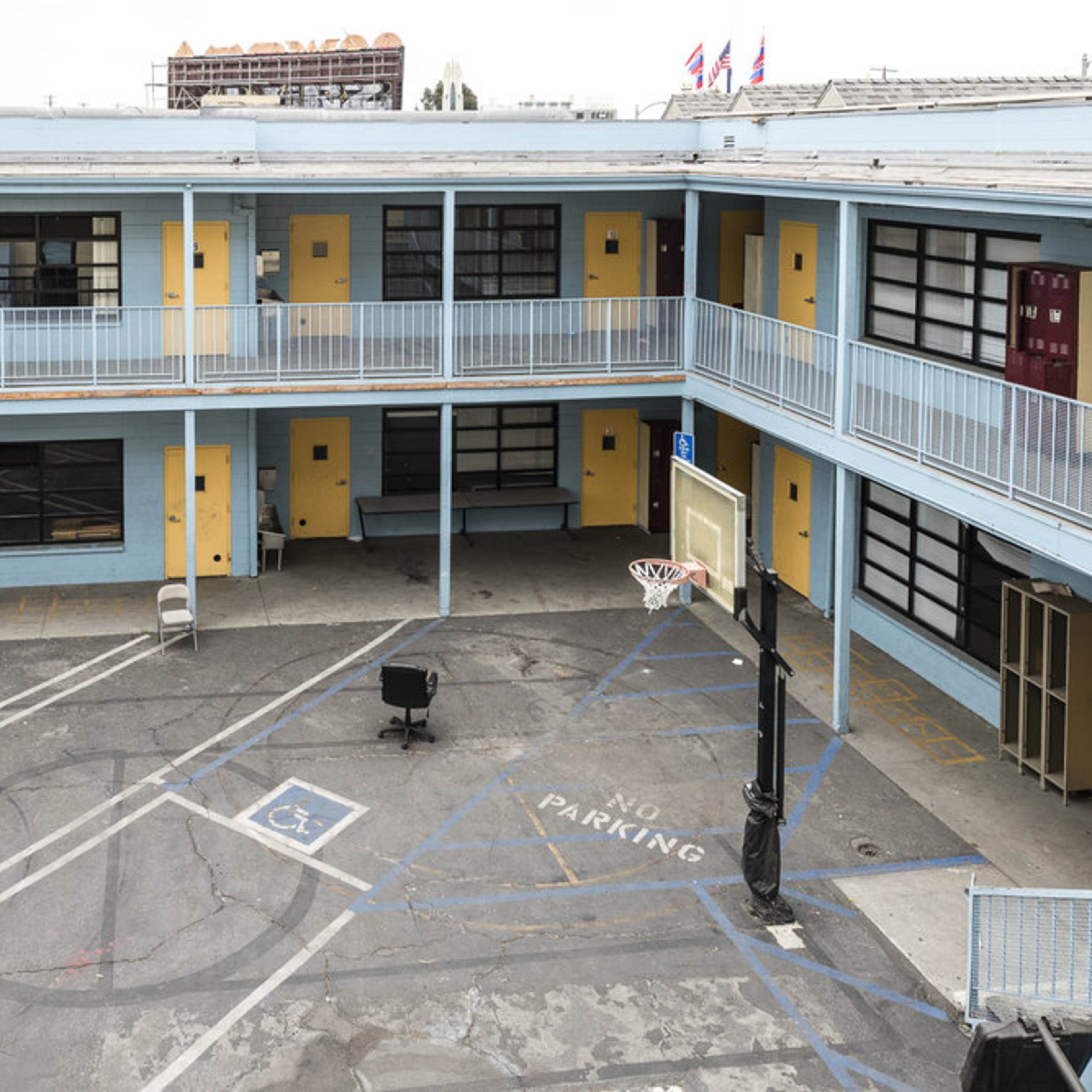 A two-story motel courtyard with rooms in yellow and blue, featuring a basketball hoop in the center and a few chairs arranged around.