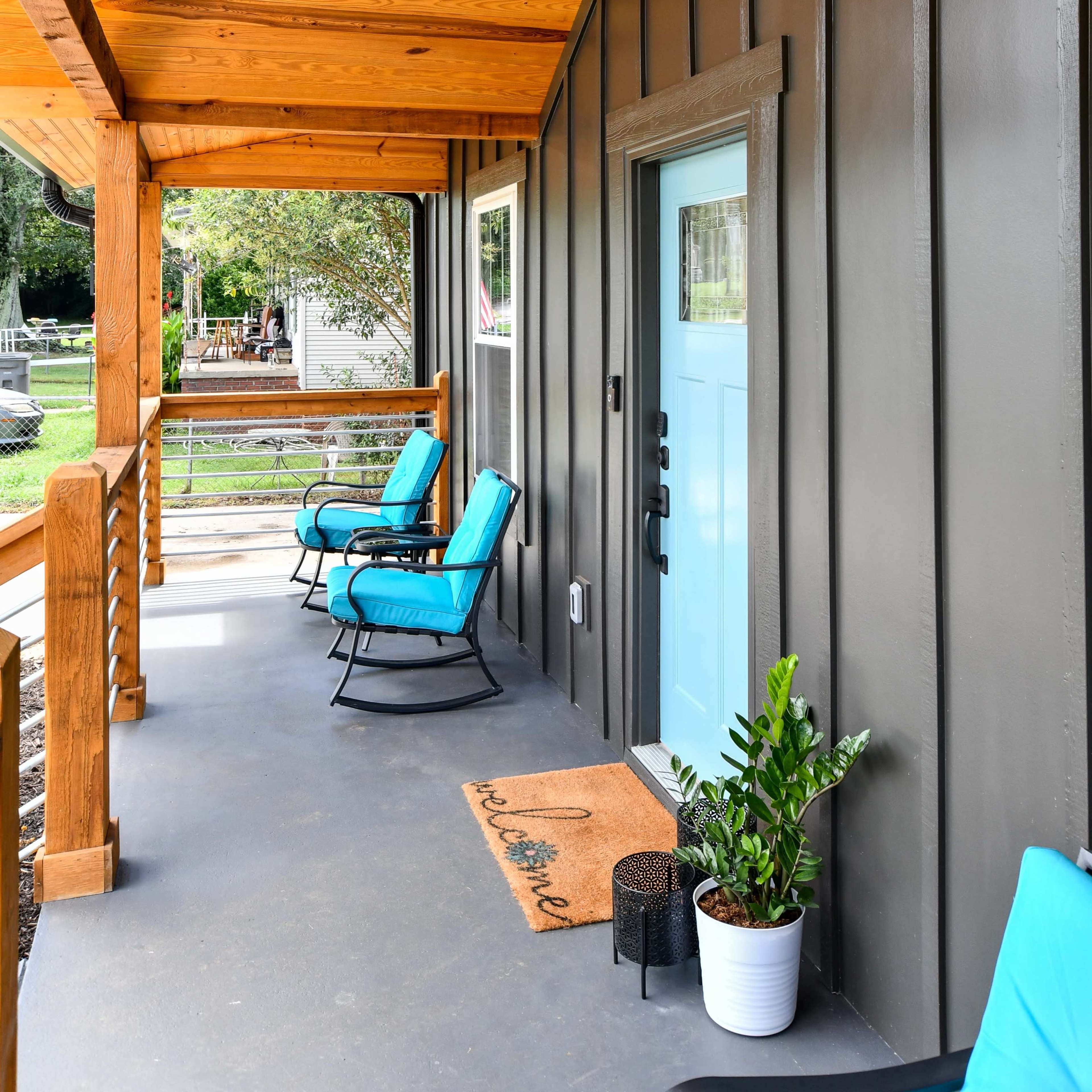 A porch with wooden ceiling beams, blue chairs, a light fixture, a light blue door, and a potted plant beside a welcome mat.