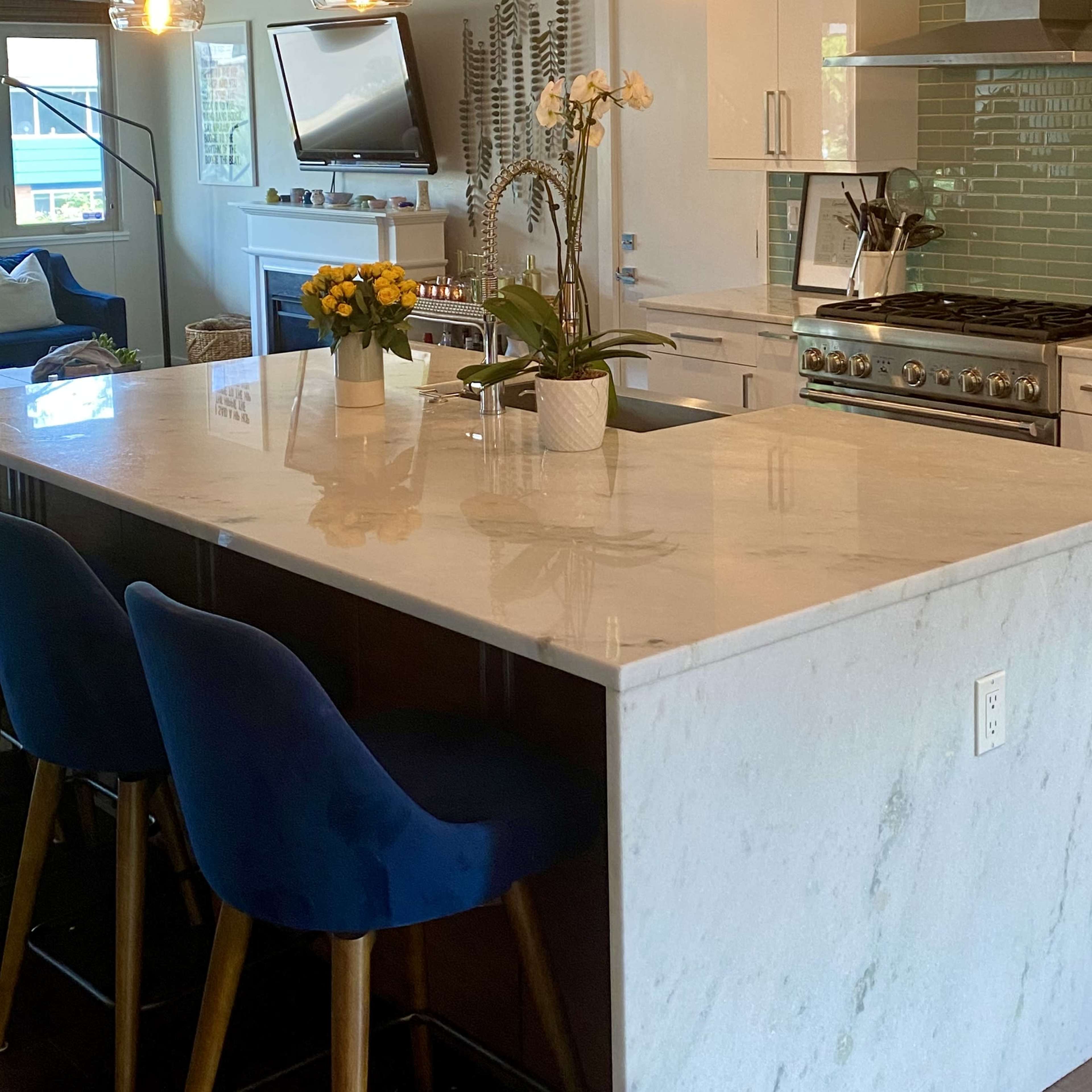 A modern kitchen features a large island with a white countertop, blue bar stools, and stainless steel appliances, surrounded by green tile and natural light.