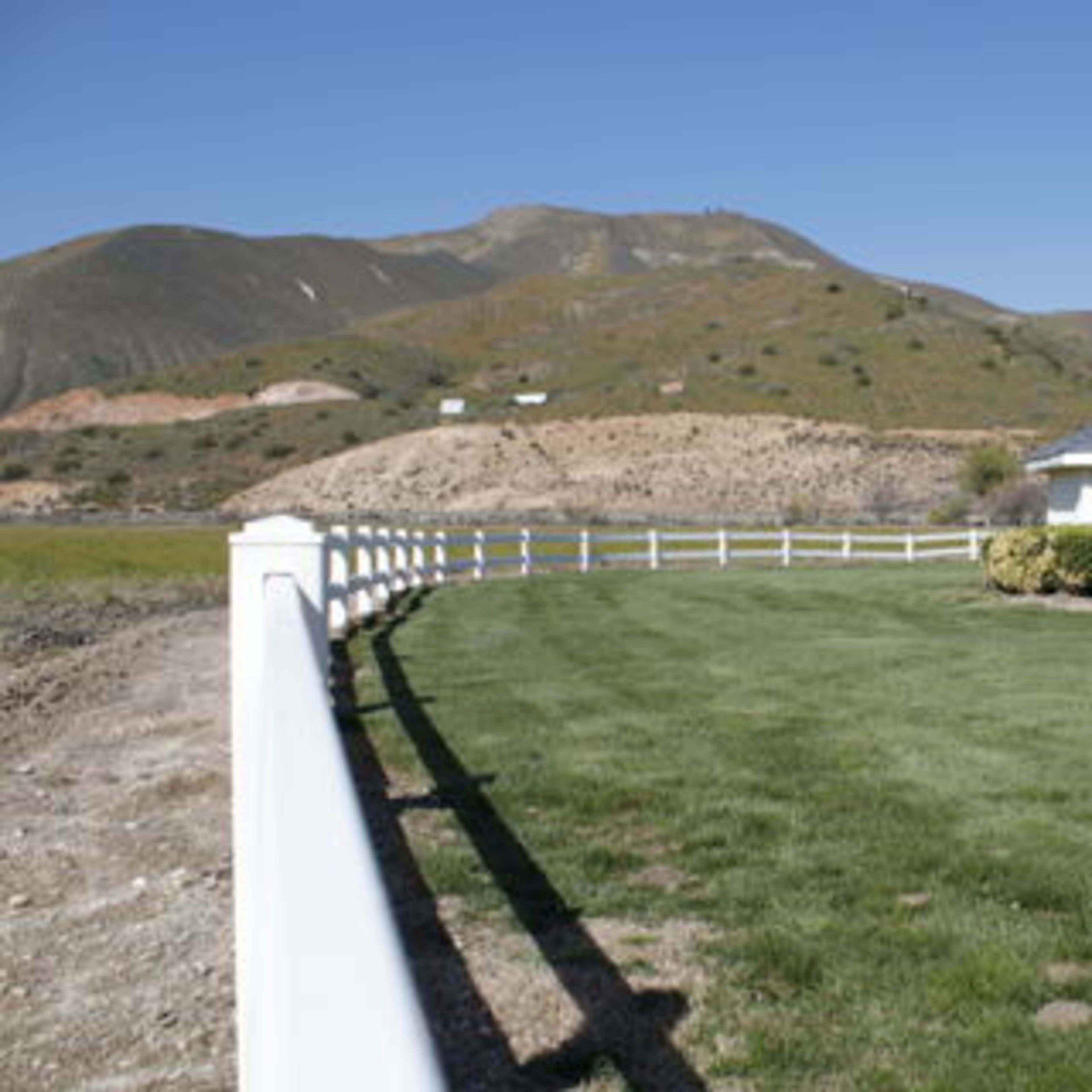 A white fence surrounds a well-maintained grassy area featuring two small buildings near a hill in the background.