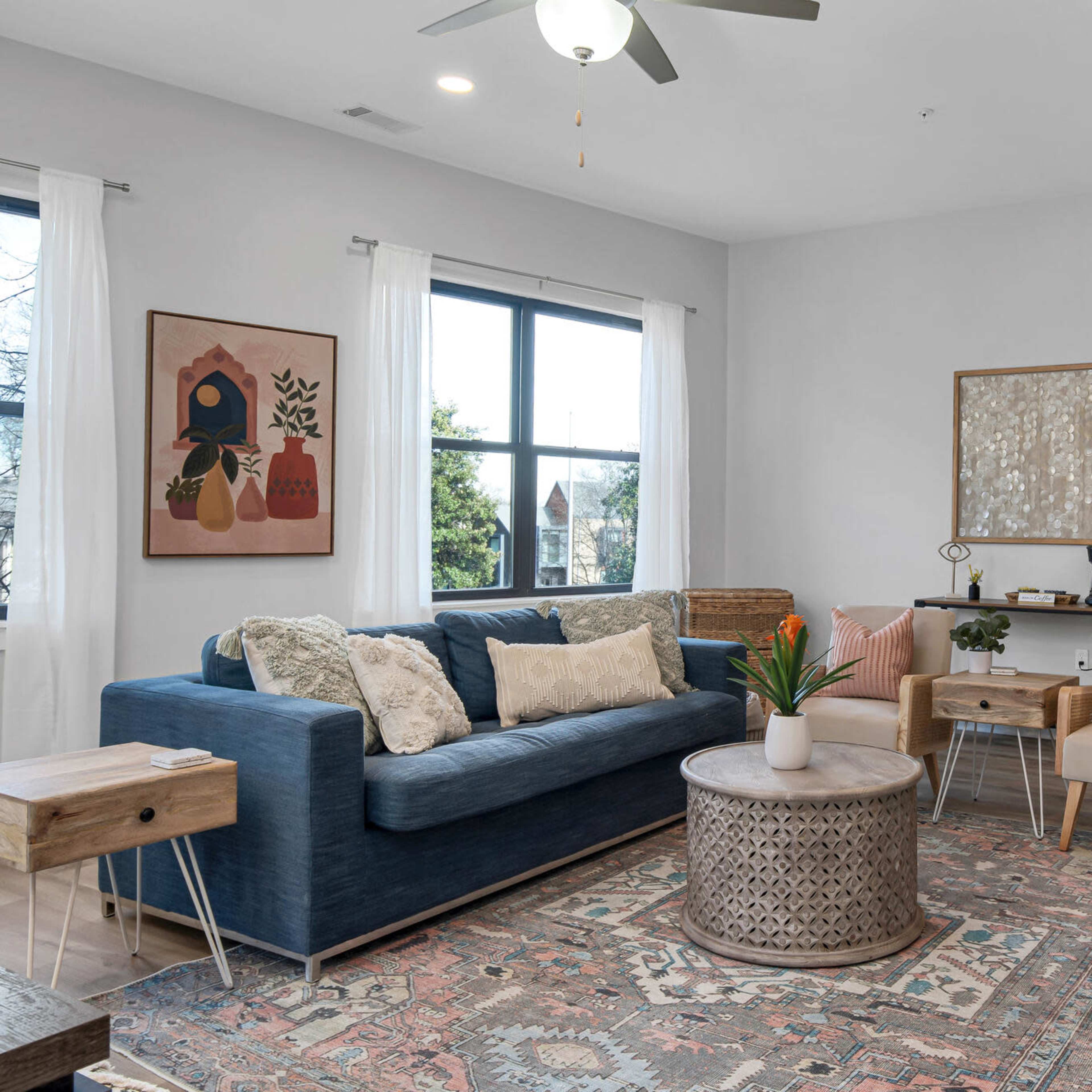 A modern living room featuring a blue sofa, wooden coffee table, decorative rug, and two chairs arranged around a stylish coffee table, with large windows allowing natural light.