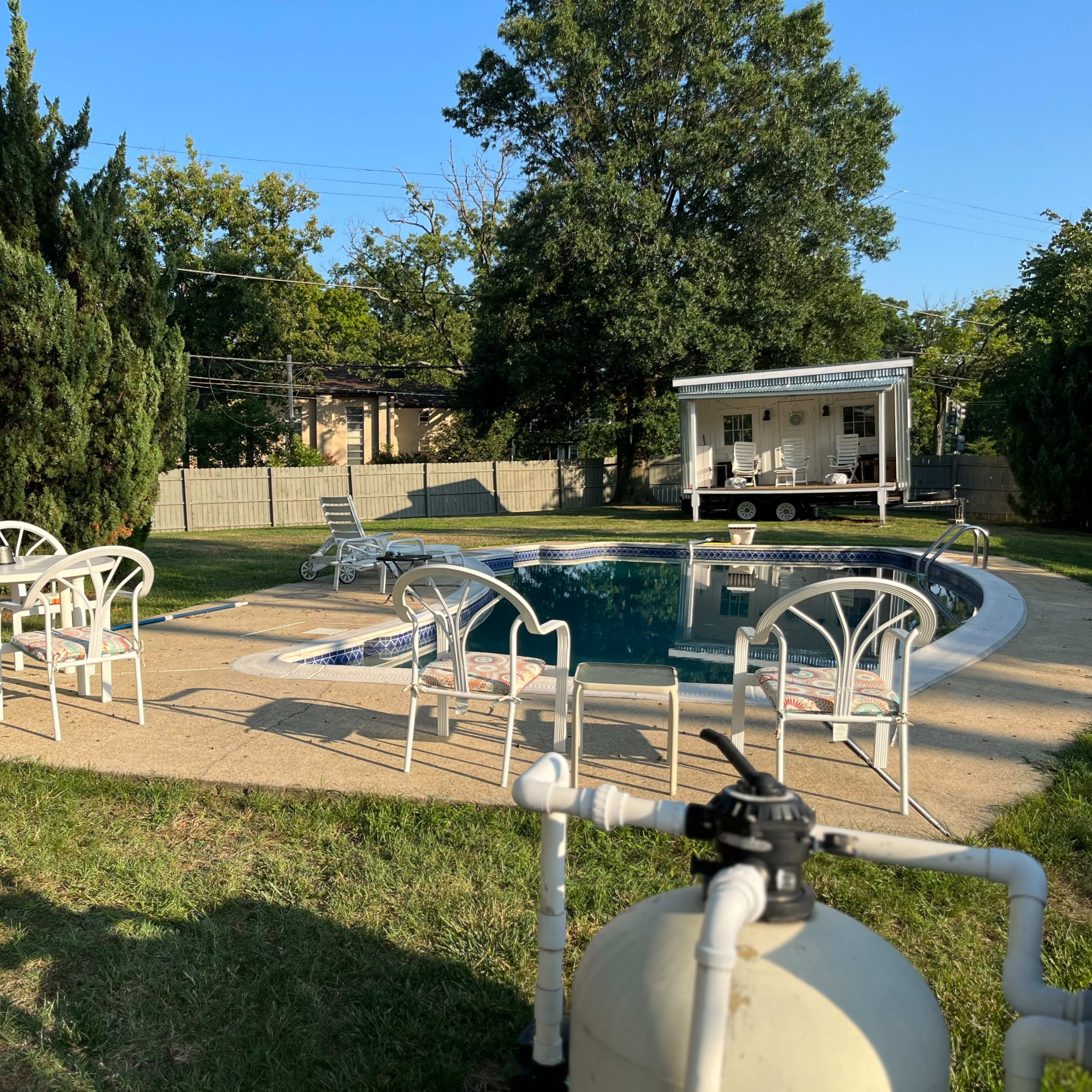 A backyard pool area features a pool, lounge chairs, a table set, and a small building in the background surrounded by trees.
