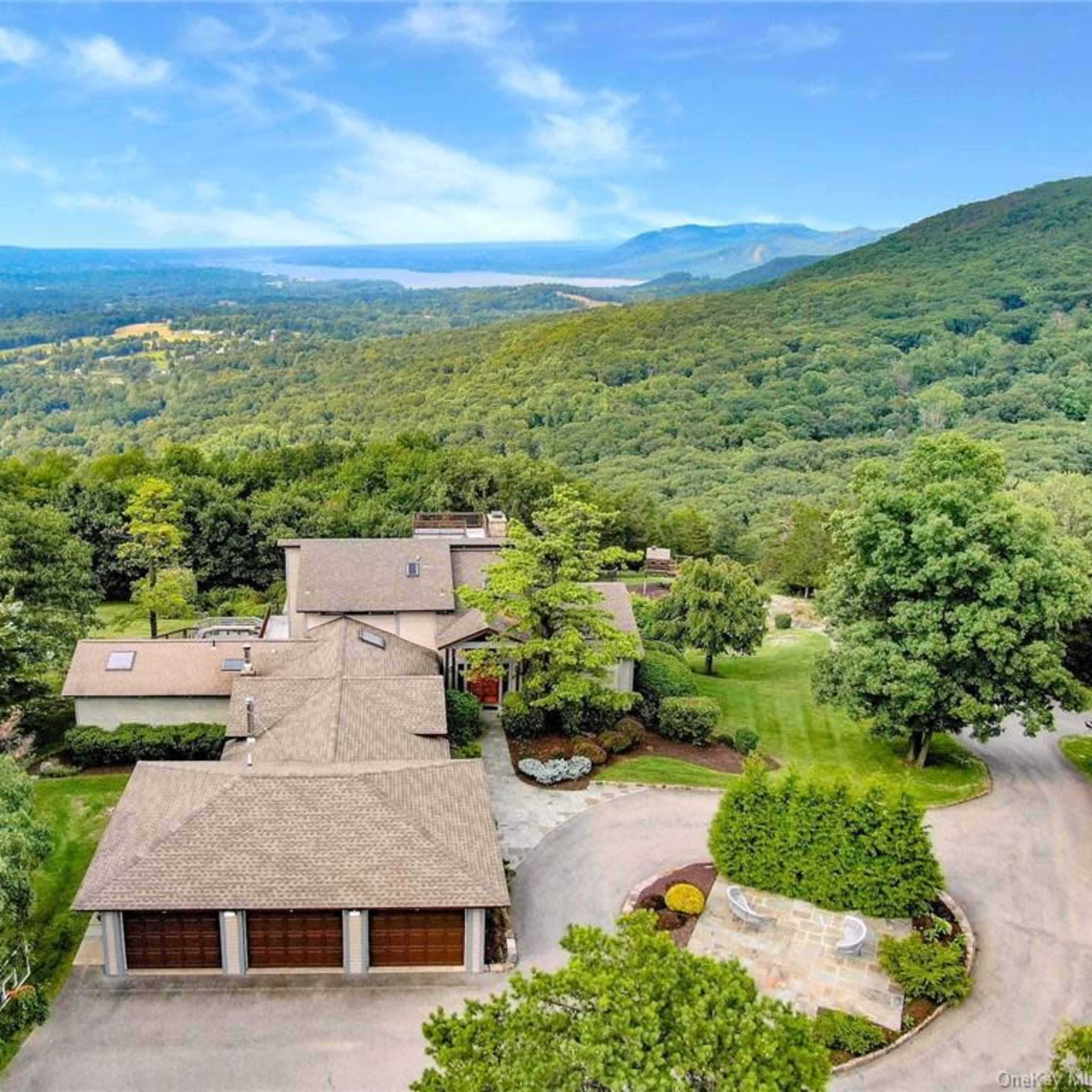 An aerial view of a large, modern house surrounded by trees and a rolling landscape, with mountains in the background.