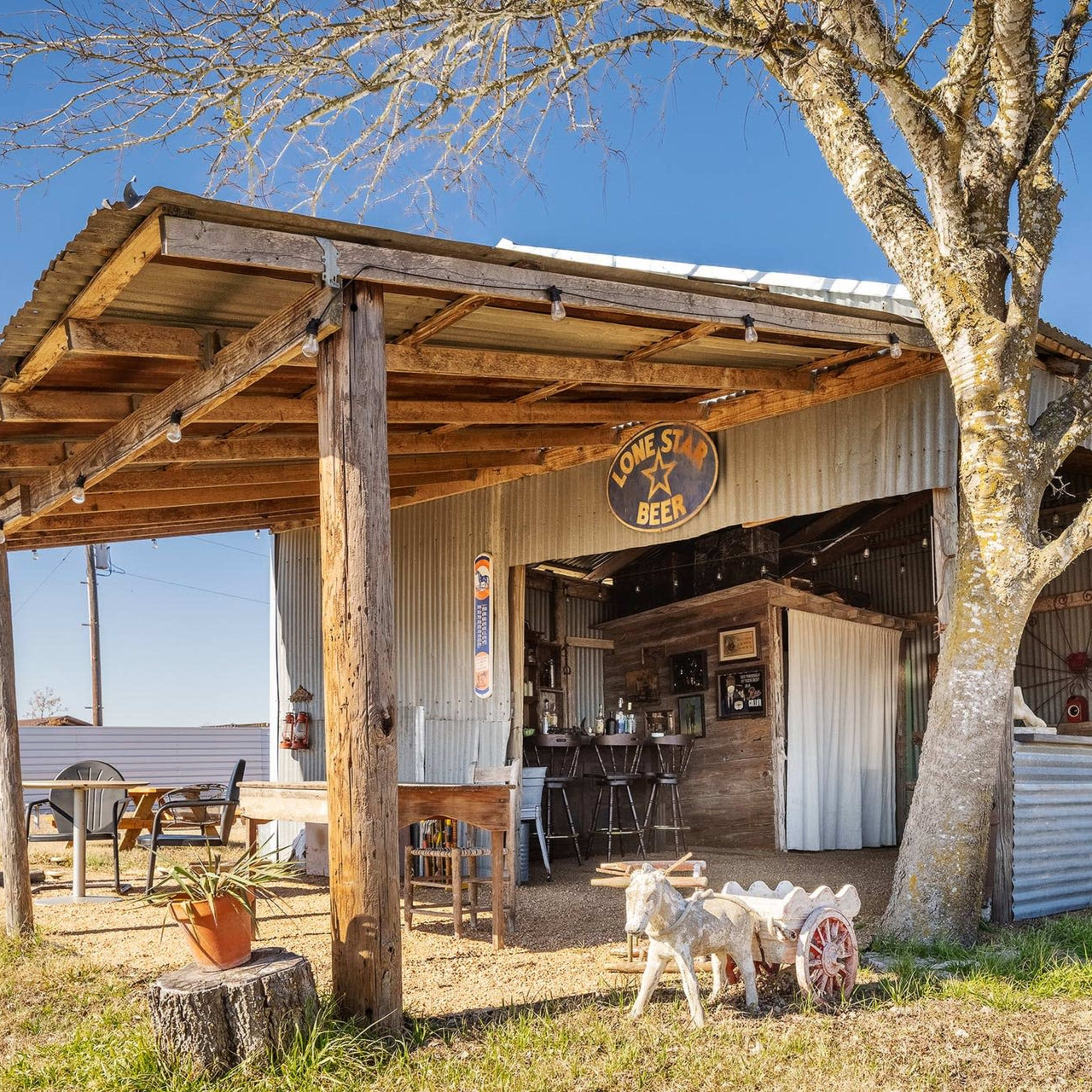 A rustic wooden shed with a tin roof and a porch features a sign that reads "Lone Star Beer" and includes outdoor seating with potted plants and a decorative cart.
