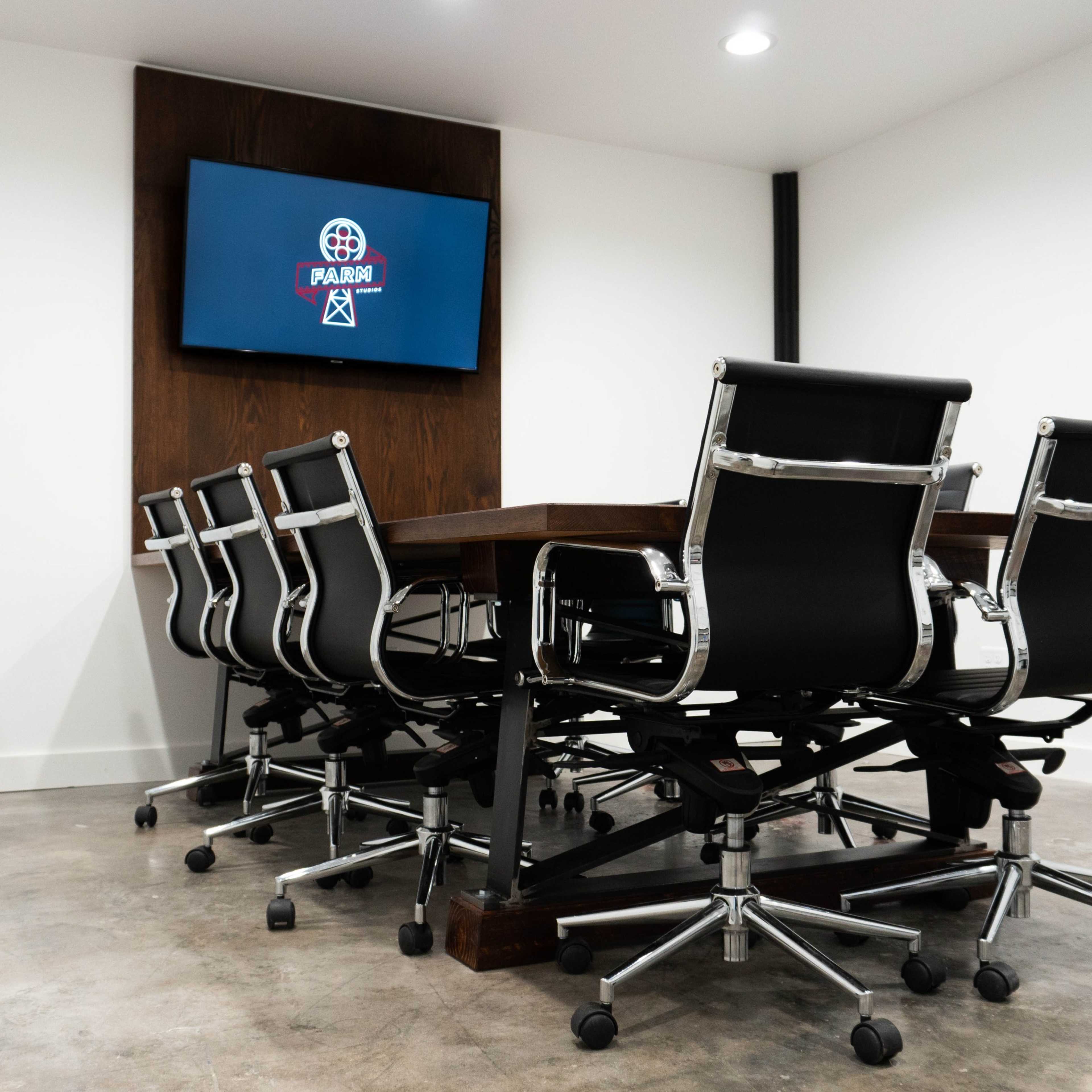 A conference room features a large wooden table surrounded by eight black rolling chairs, with a television mounted on the wall displaying a logo.