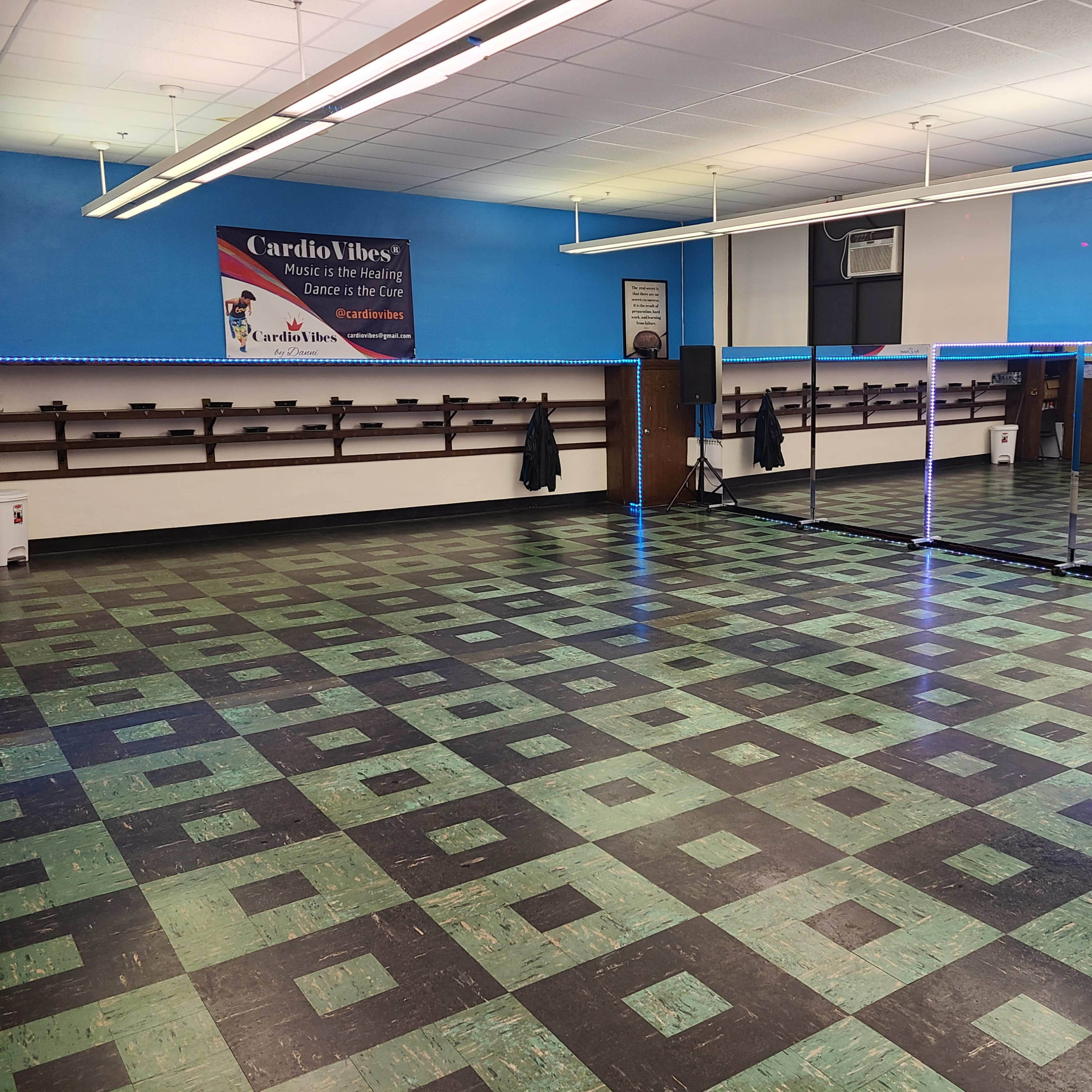 A spacious dance studio featuring a checkerboard floor, mirrored walls, and shelves along one side.