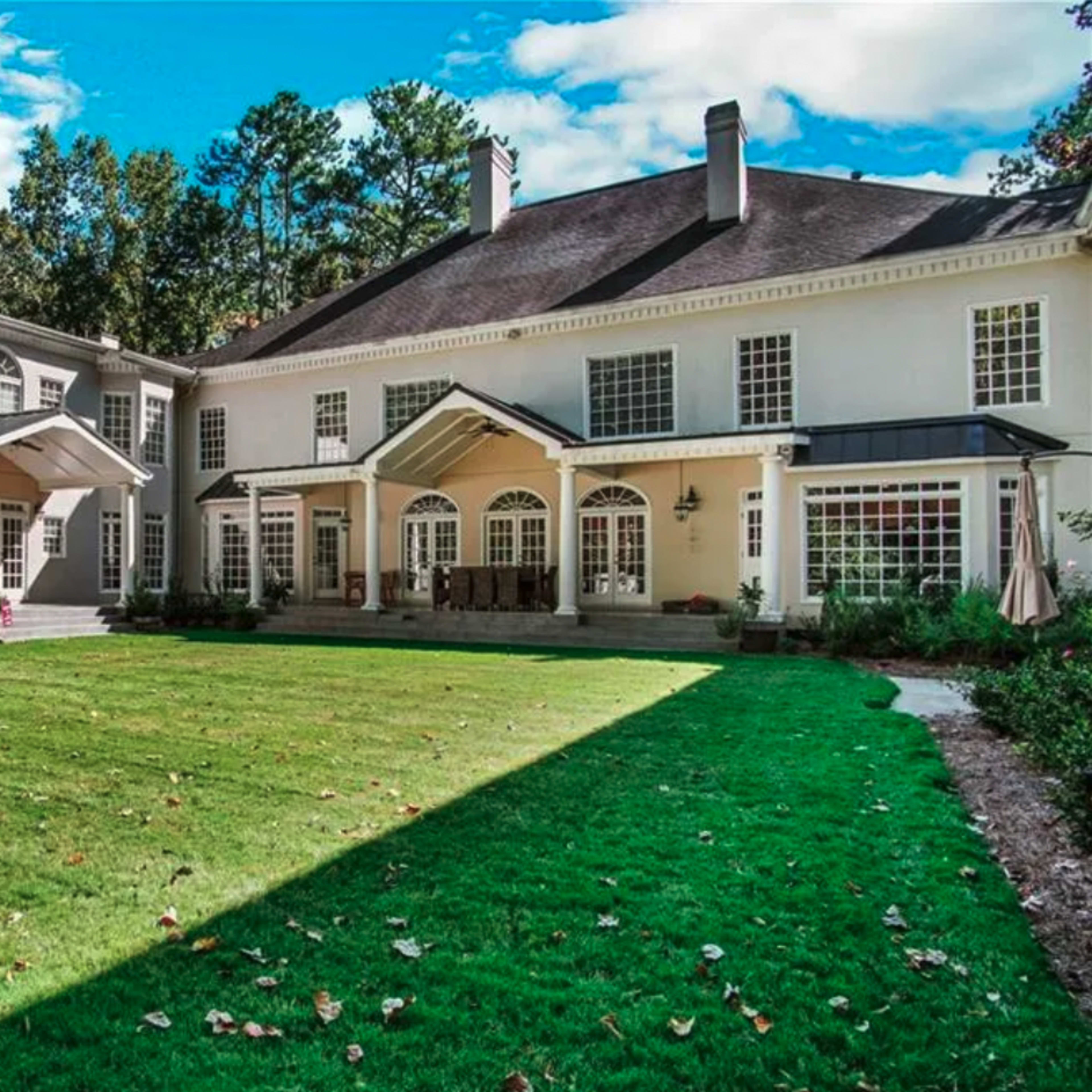 A large two-story home with multiple gabled roofs, a spacious lawn, and a patio area surrounded by trees.
