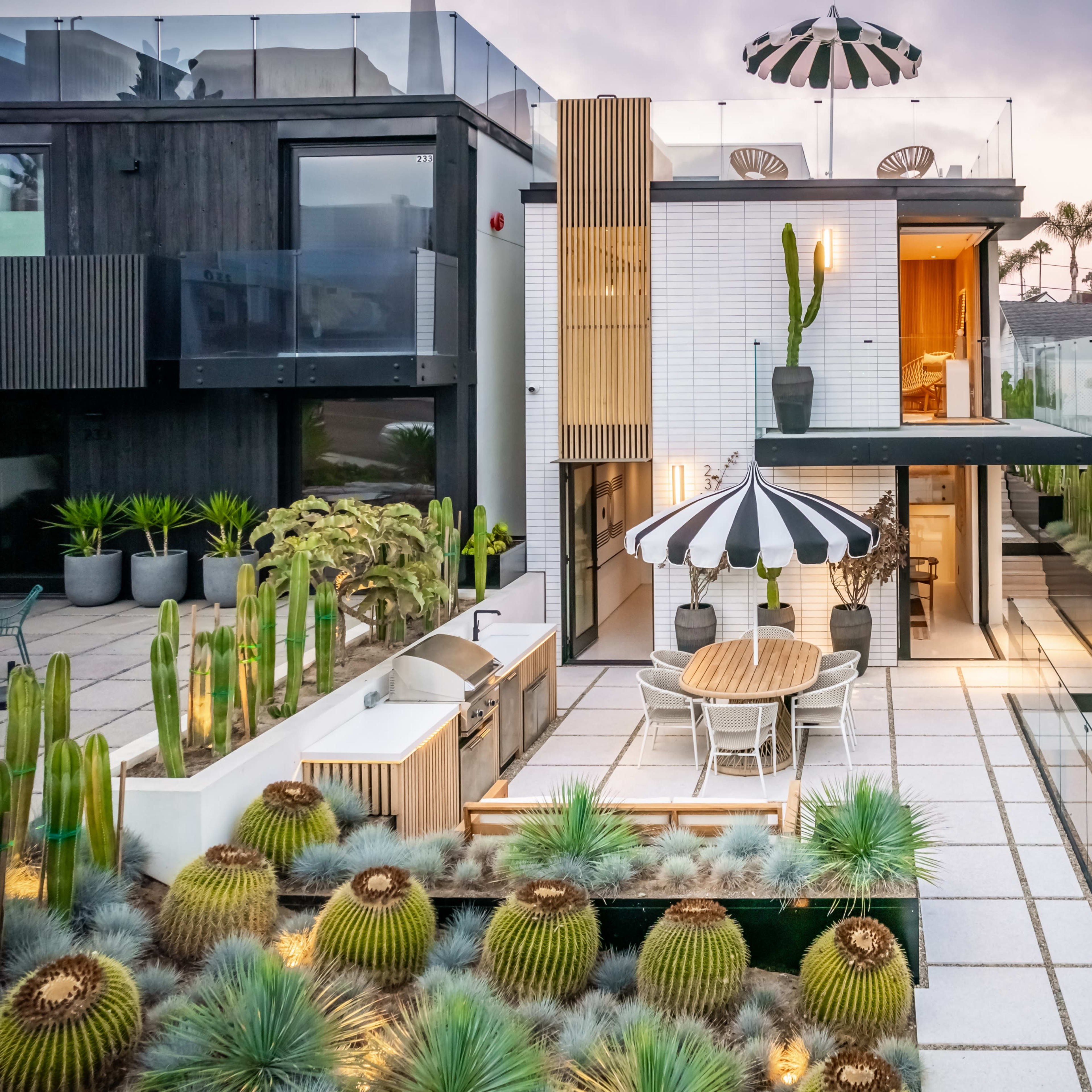 A modern house with a rooftop terrace featuring a striped umbrella, surrounded by a landscaped garden filled with cacti and a dining area.