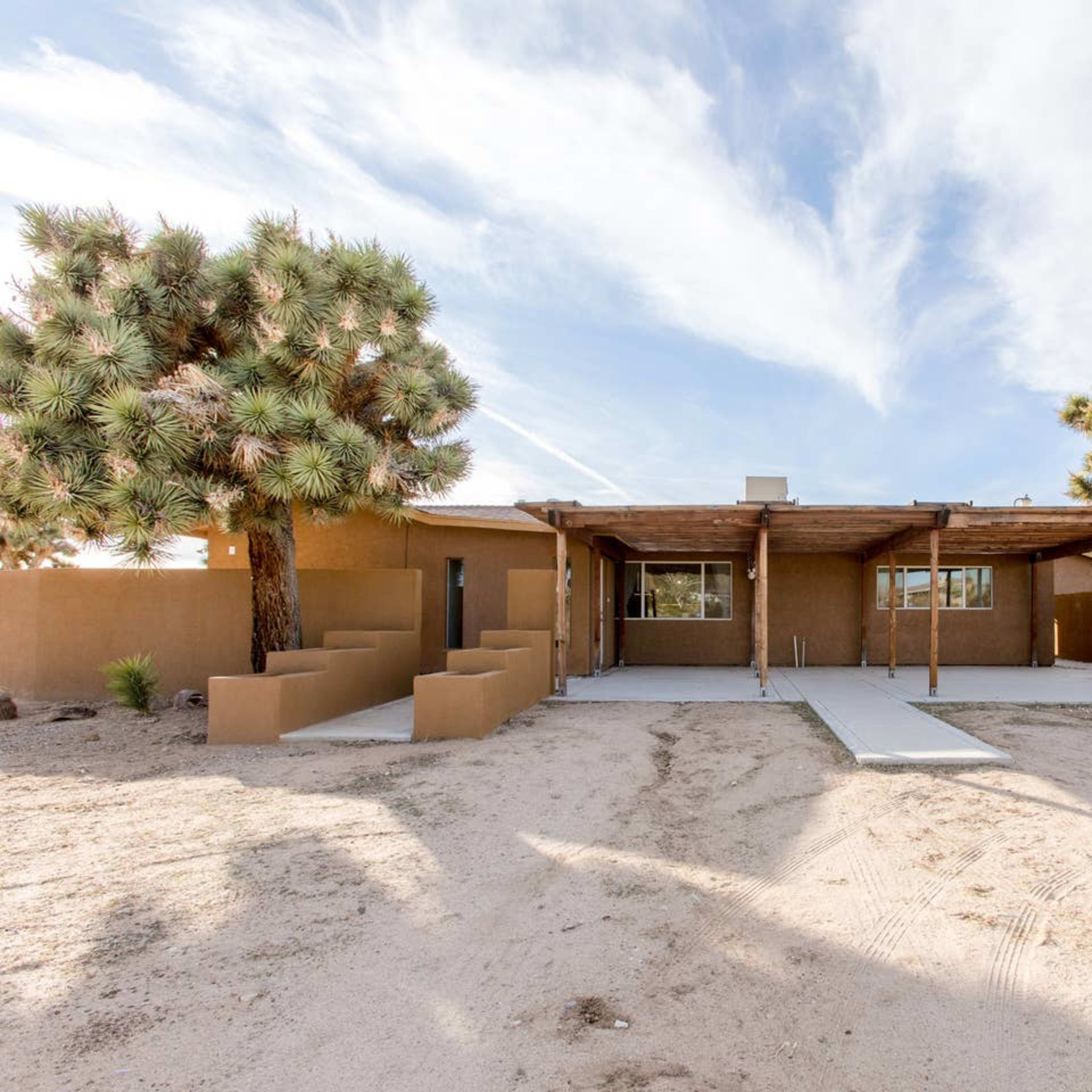 A single-story brown house with a covered porch, surrounded by desert landscape and Joshua trees.