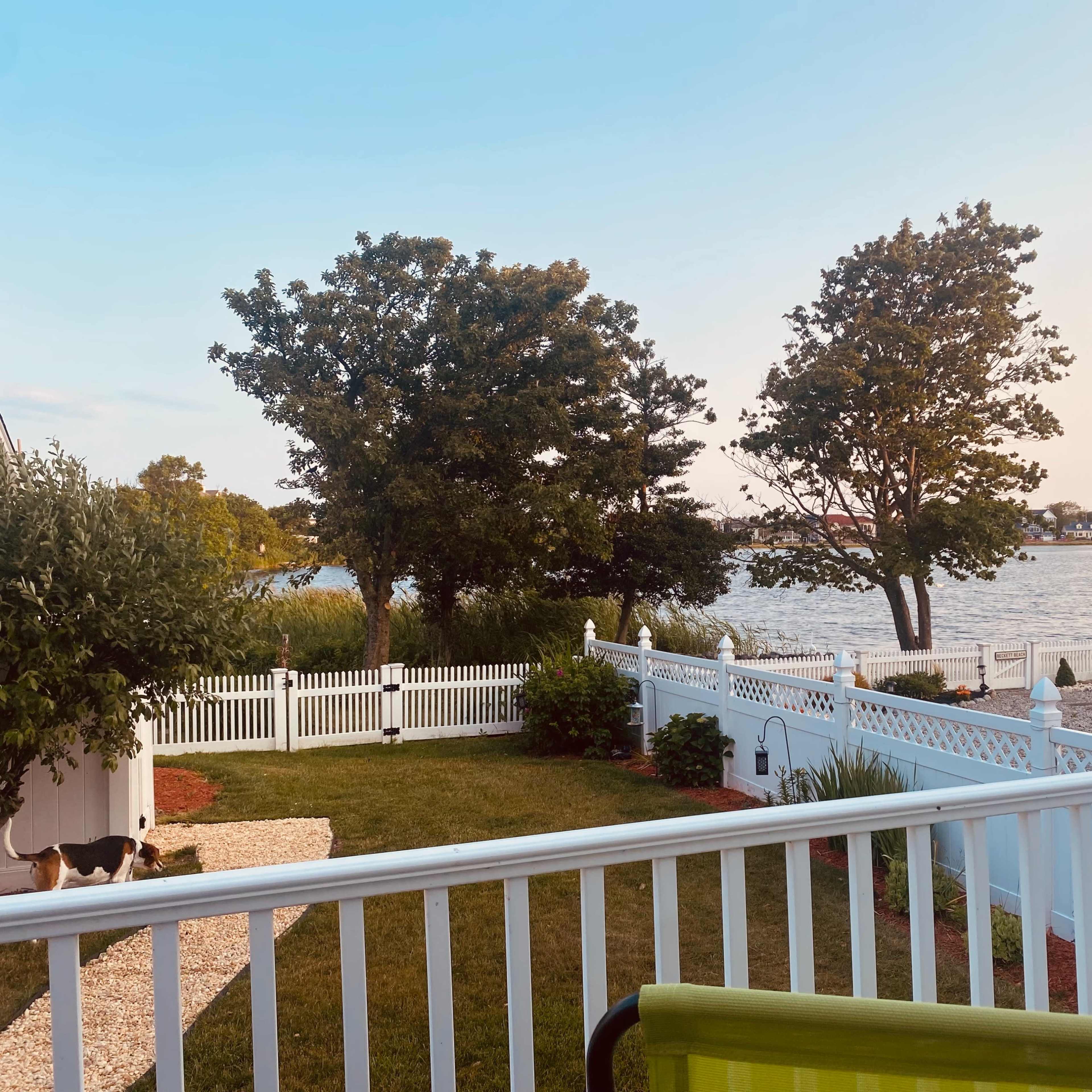 A view of a waterfront area with trees and houses in the distance, framed by a white picket fence and lawn chairs on the patio.