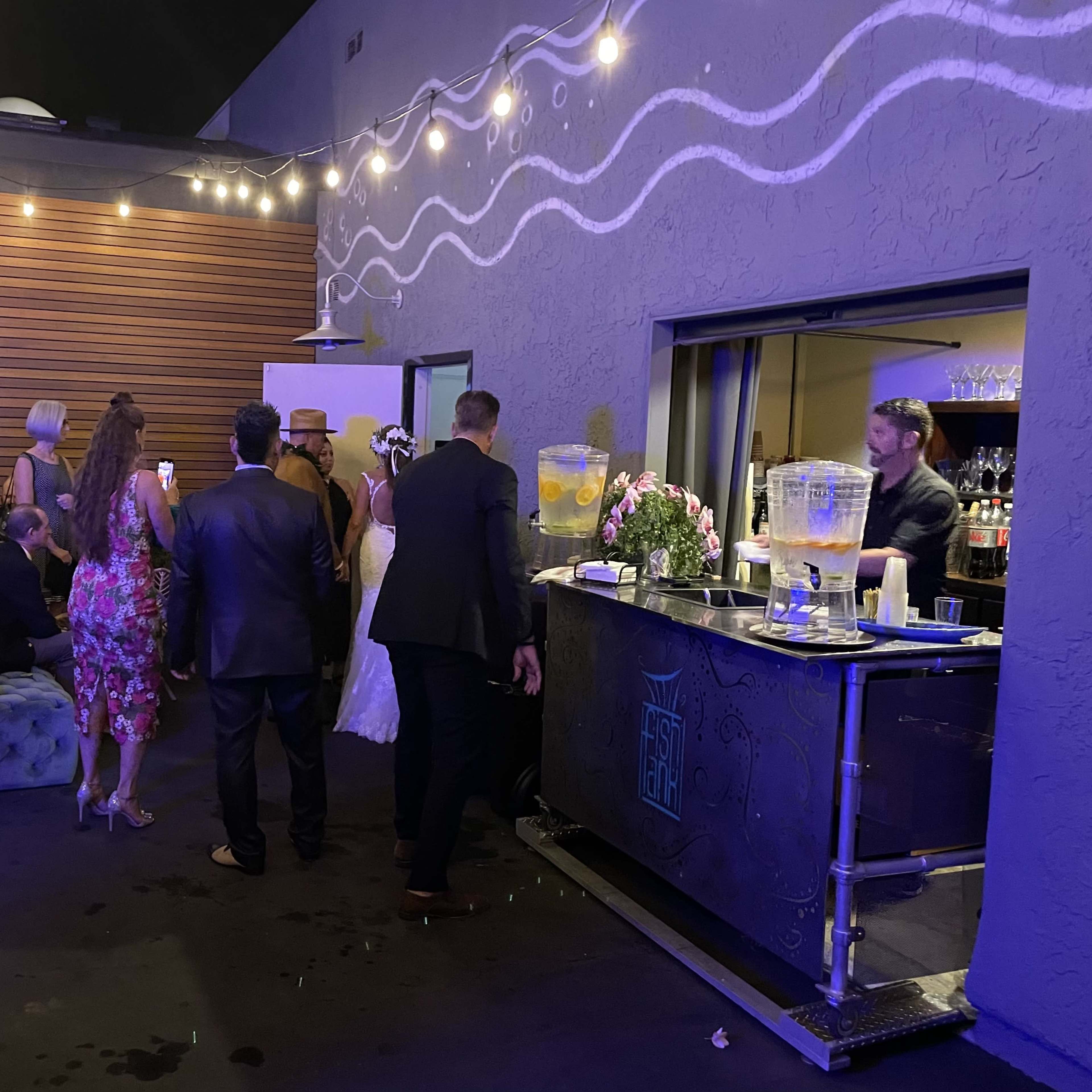 A group of people in formal attire gather around a bar area with drinks and decorations, under string lights.