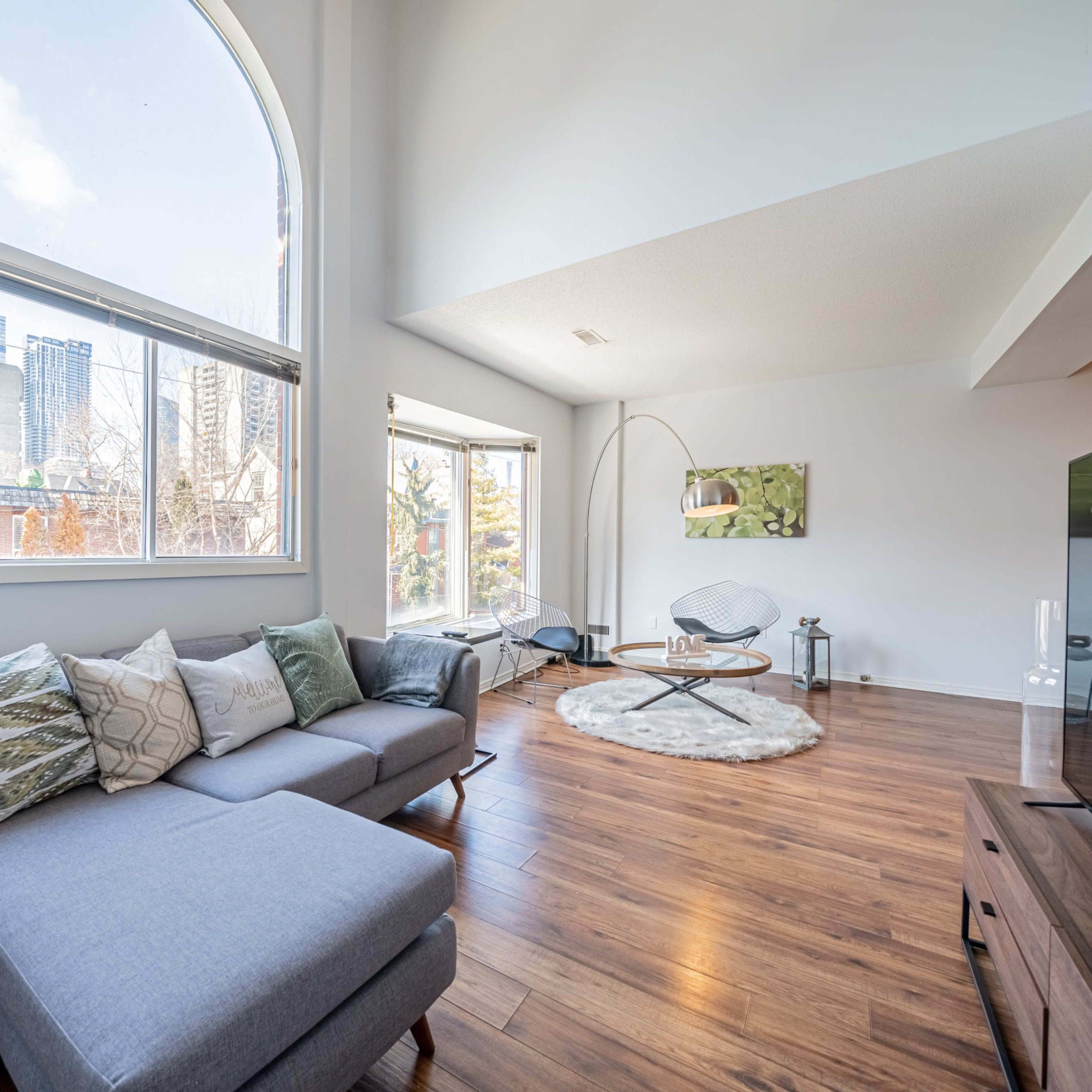 A modern living room features a gray sectional sofa, a round rug, and large windows letting in natural light, with city skyline views in the background.